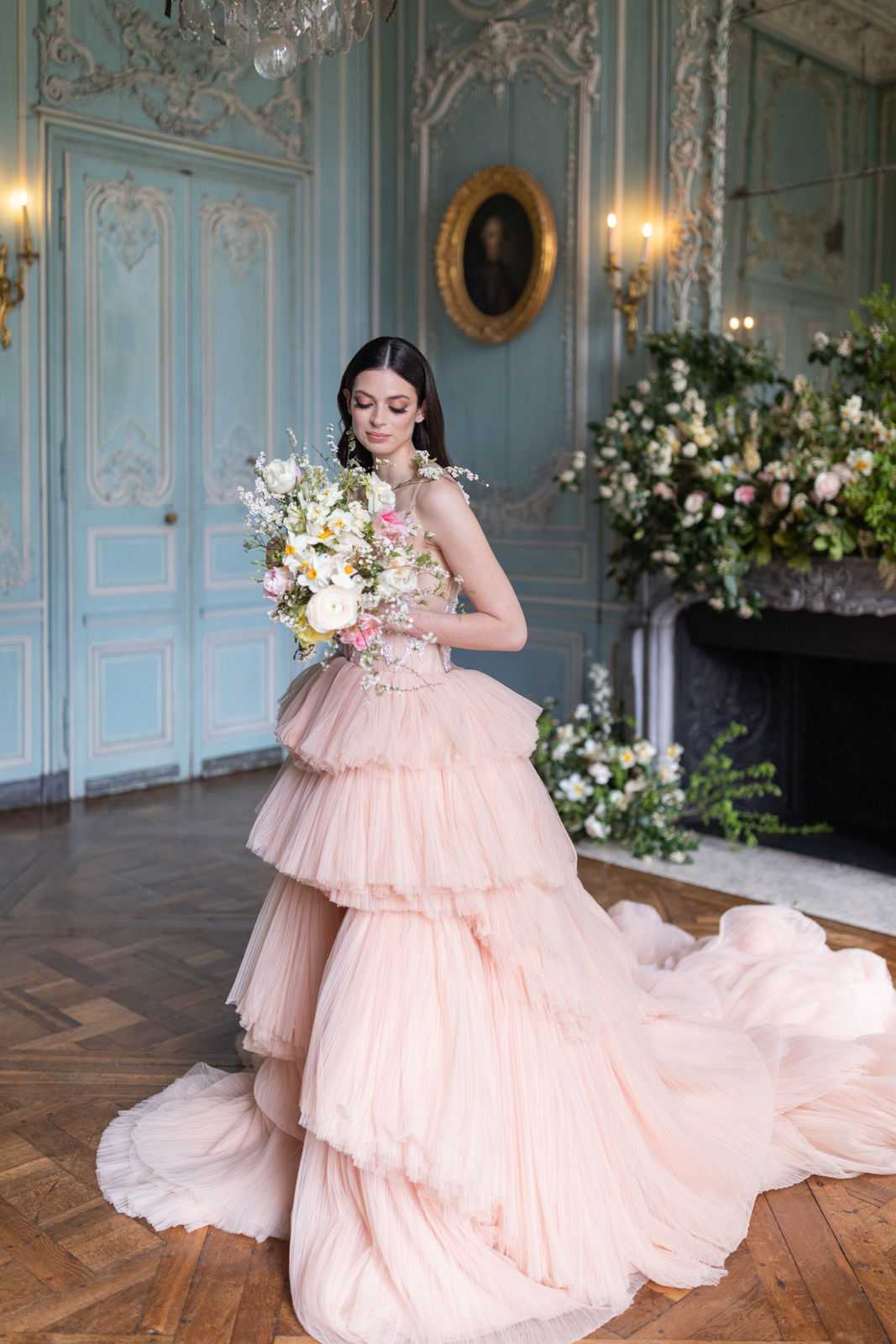 A bridal portrait shot indoors in a French chateau salon, featuring a bride in a blush pink tiered tulle ball gown with a dramatic train and a corset-style bodice with floral appliqué details. She holds a loose, garden-style bouquet composed of white ranunculus, cream tulips, yellow daffodils, hot pink blooms, and delicate white filler flowers, and is looking downward toward the bouquet. The room features pale blue-grey boiserie paneling with white and gilt rococo plasterwork, a crystal chandelier, a marble fireplace flanked by candle wall sconces, and an oval gilt-framed portrait on the wall. A large lush floral installation in shades of blush, white, and yellow with abundant greenery overflows the fireplace mantel and spills onto the floor. The herringbone parquet floor and the overall French rococo interior point to a classic chateau setting styled with a romantic, maximalist floral approach. Medium full-length portrait composition. Potential venue feature image.