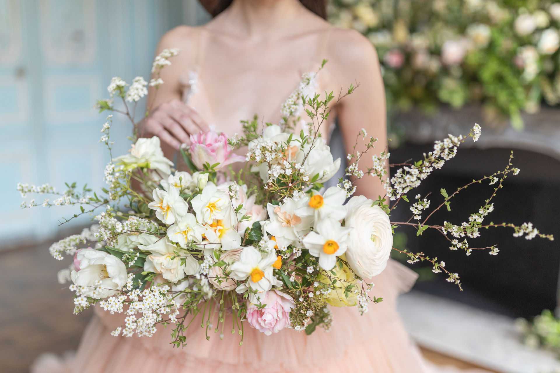 A close-up portrait shot of a bride holding a large, loosely arranged bridal bouquet composed of white narcissi with yellow-orange centers, white ranunculus, pink tulips, blush garden roses, and trailing branches of white blossom with fresh green foliage. The bride is wearing a blush peach tulle gown with thin spaghetti straps, and the image is cropped at the neck, keeping focus on the bouquet. The setting appears to be indoors near a fireplace, with a soft blue-grey wall visible in the background and a large floral arrangement of blush pink and white blooms on the mantelpiece behind her. The overall styling palette is soft spring — blush, white, and pale yellow — with a romantic, garden-gathered aesthetic.
