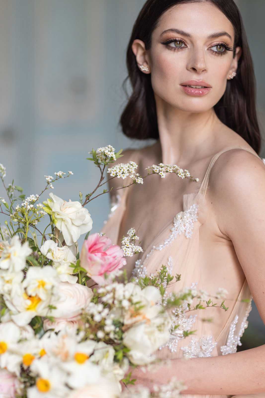 A close-up portrait of a bride holding a large, loosely arranged bouquet featuring blush pink roses, white blossom branches, yellow-centered white daisies, and soft pink peonies with green foliage. The bride wears a blush-toned gown with sheer tulle and white beaded lace appliqué detailing, thin spaghetti straps, and small pearl or crystal cluster stud earrings. Her makeup includes defined winged liner, full lashes, and a nude-pink lip. Her dark hair is worn down in a soft, slightly wavy style. The composition is a tight portrait shot with a soft, muted blue-grey background, giving the image a modern, editorial feel.