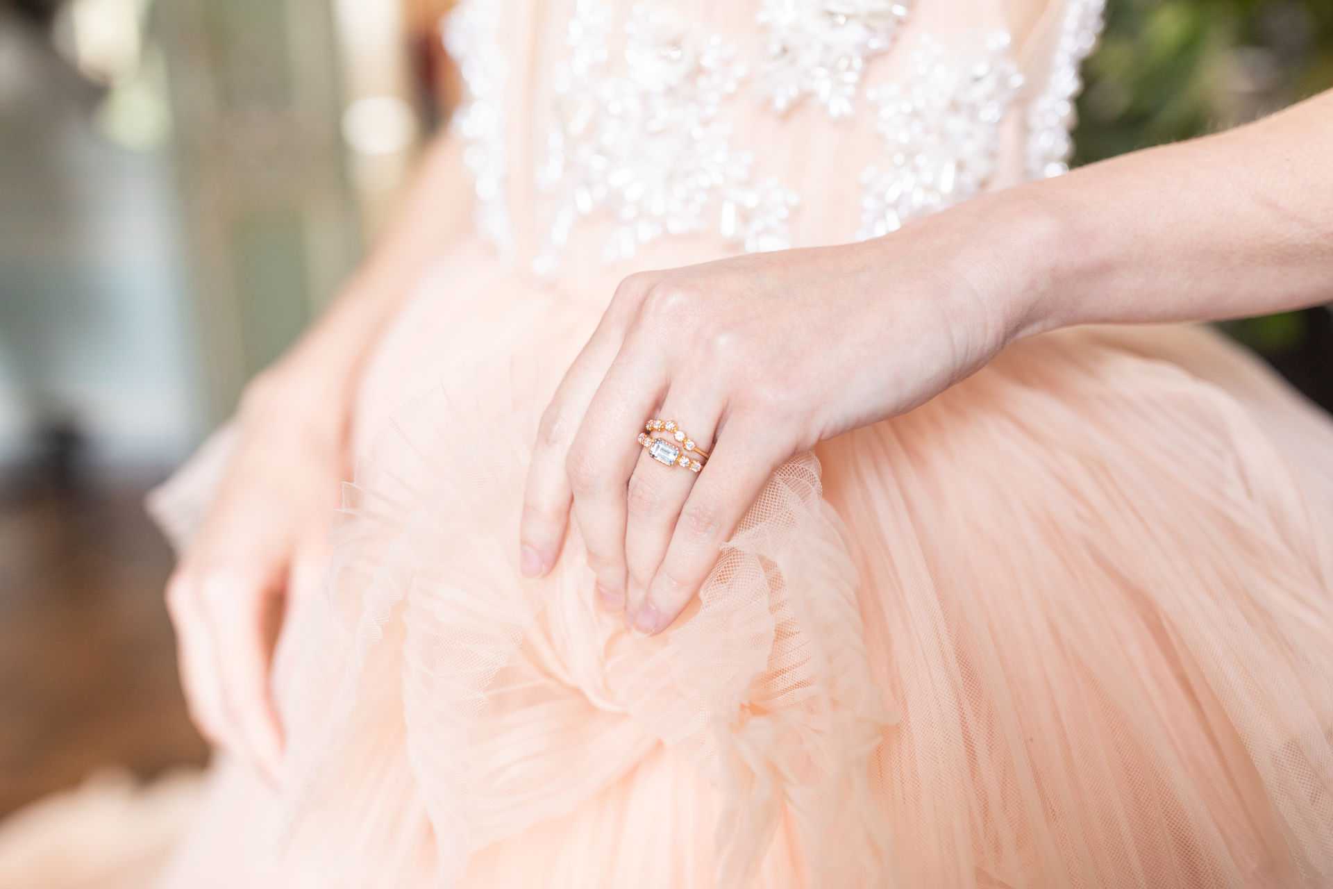 A close-up detail shot of a bride's hands resting on the skirt of a blush peach tulle ballgown with a heavily beaded and crystal-embellished bodice visible at the top of the frame. The bride wears a rose gold engagement ring featuring an emerald-cut center stone flanked by round diamonds, stacked with a matching rose gold diamond wedding band. The soft tulle fabric of the skirt fills most of the frame, emphasizing the ring and dress fabric as the focal point of the composition.