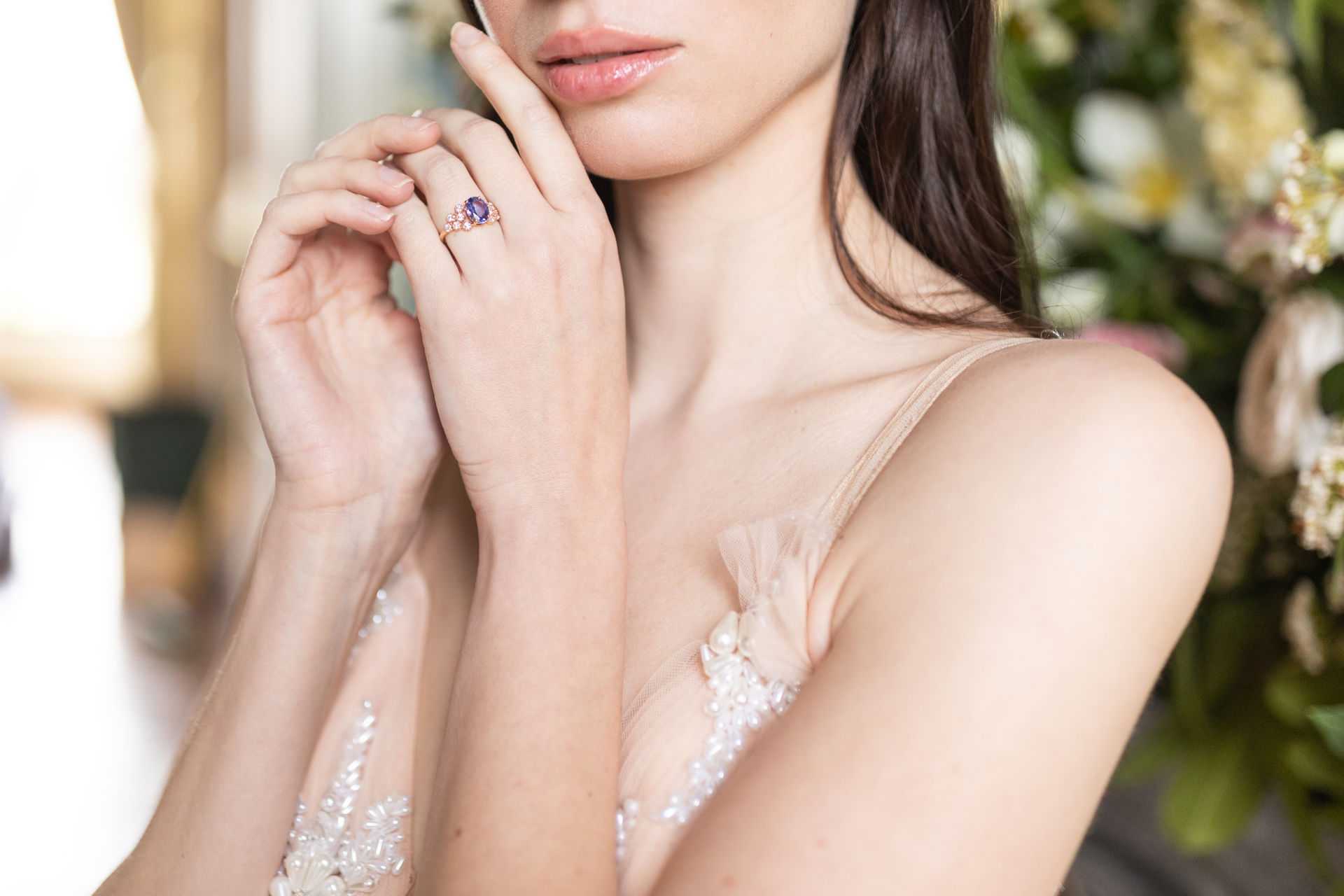 A close-up portrait detail shot of a bride, framing her from the chin to the chest with her hand raised near her face. The focal point is a rose gold engagement ring featuring a central oval purple/violet gemstone surrounded by small pavé diamonds. She is wearing a blush-toned gown with delicate sheer straps adorned with pearl and beaded floral appliqué. The background is softly blurred, showing what appears to be cream and blush florals, greenery, and a gilded mirror frame, suggesting an indoor getting-ready setting with classic French interior decor.