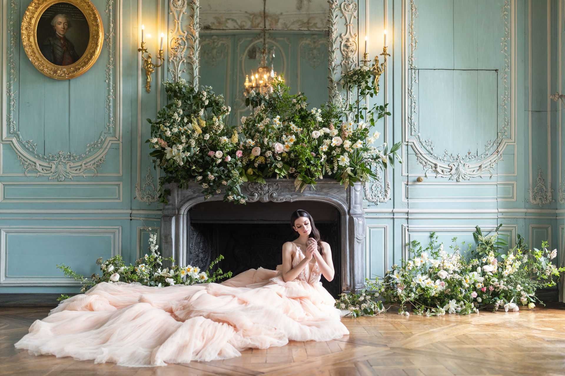 A bridal editorial portrait shot indoors in what appears to be a French chateau salon, featuring a bride seated on a herringbone parquet floor in front of an ornate carved stone fireplace. The bride wears a voluminous blush pink tulle ballgown with a lace bodice, her dark hair pulled back, and she looks downward with hands clasped. The room features pale duck-egg blue boiserie paneling with white carved moldings, gold candle wall sconces, a small crystal chandelier visible in the mirror above the mantel, and an oil portrait in a gilded frame on the left wall. The fireplace mantel and floor on both sides are covered in an abundant, loose floral installation featuring white narcissus, blush garden roses, ranunculus, white lisianthus, soft yellow blooms, and trailing greenery including ferns and leafy vines. The overall decor palette combines blush, white, cream, and green against the blue-grey interior. Wide portrait shot. Potential venue feature image.
