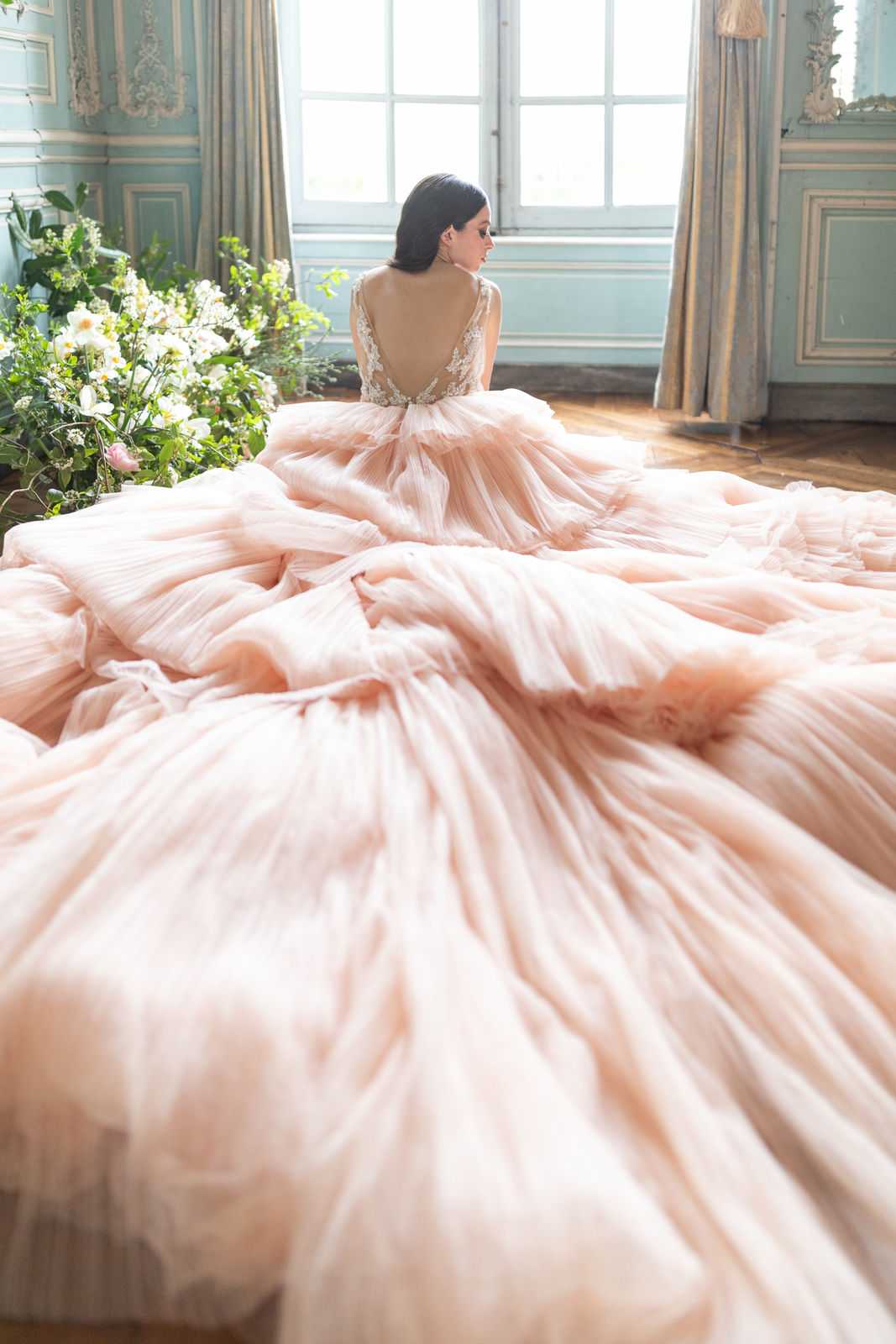 A bridal portrait shot from behind inside a French chateau salon, showcasing a bride wearing a voluminous blush pink tulle ball gown with an extremely long, ruffled train that spreads across the entire herringbone parquet floor. The bodice features a deep V-back neckline with lace and floral appliqué detailing. The bride, who has dark hair worn up, stands facing a large multi-pane window that floods the room with natural light. To her left, a large loose floral arrangement sits on the floor featuring white blooms, pink roses, and abundant greenery. The room's walls are painted pale blue-grey with white ornate boiserie paneling, consistent with classical French chateau interiors. The composition is a wide portrait taken from floor level, emphasizing the scale of the train in the foreground while the bride and room recede toward the window in the background. Potential venue feature image.