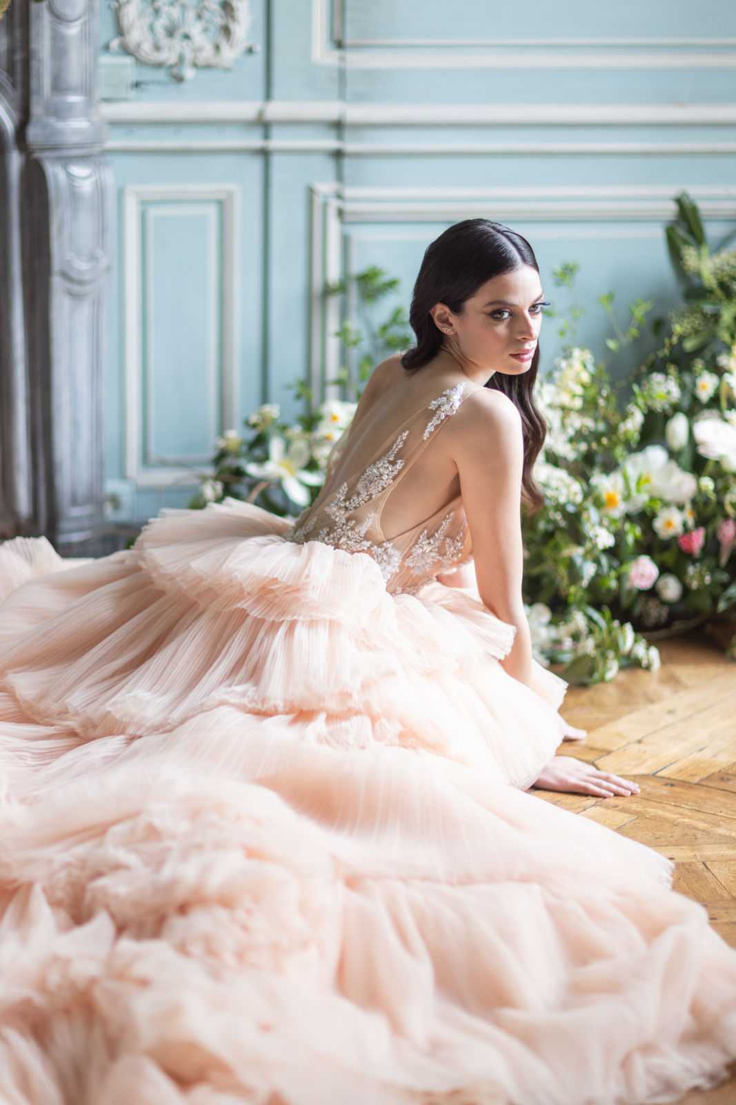 A bridal portrait of a single dark-haired bride seated on a herringbone parquet wood floor, looking back over her shoulder toward the camera. She wears a voluminous blush pink tiered tulle ball gown with a sheer nude illusion bodice featuring white floral lace appliqué and a deep open back. The full skirt spreads widely across the floor in layered pleated tiers. The indoor setting features pale blue-grey panelled boiserie walls with ornate white plasterwork molding and a carved fireplace surround, consistent with a French chateau or hôtel particulier interior. A large loose floral arrangement in the background includes white peonies, cream roses, white daffodils, pink blooms, and lush greenery placed at floor level. The styling aesthetic is classic French with romantic, editorial influences. Medium portrait shot taken from behind and slightly to the side, emphasizing the gown's back detail and train.