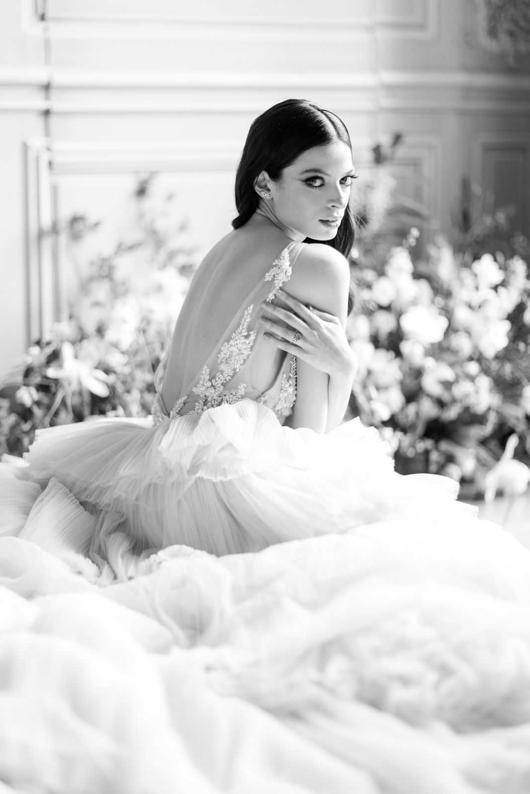 This is a black-and-white bridal portrait of a bride seated indoors, turning to look over her shoulder toward the camera. She wears a voluminous ball gown with a deep open back, sheer lace appliqué detailing along the bodice straps, and a full tiered tulle skirt that spreads widely around her. Her dark hair is pulled back sleekly, and she wears statement crystal earrings and a ring visible on her hand, which rests on her arm. The background features large lush floral arrangements with full blooms and leafy greenery, set against ornate paneled wall molding consistent with a classic French interior such as a chateau or hôtel particulier. The image is rendered in high-contrast black and white with soft highlights and deep shadows that emphasize the texture of the tulle and lace. The composition is a close-to-medium portrait shot taken from a low angle, foregrounding the skirt volume.