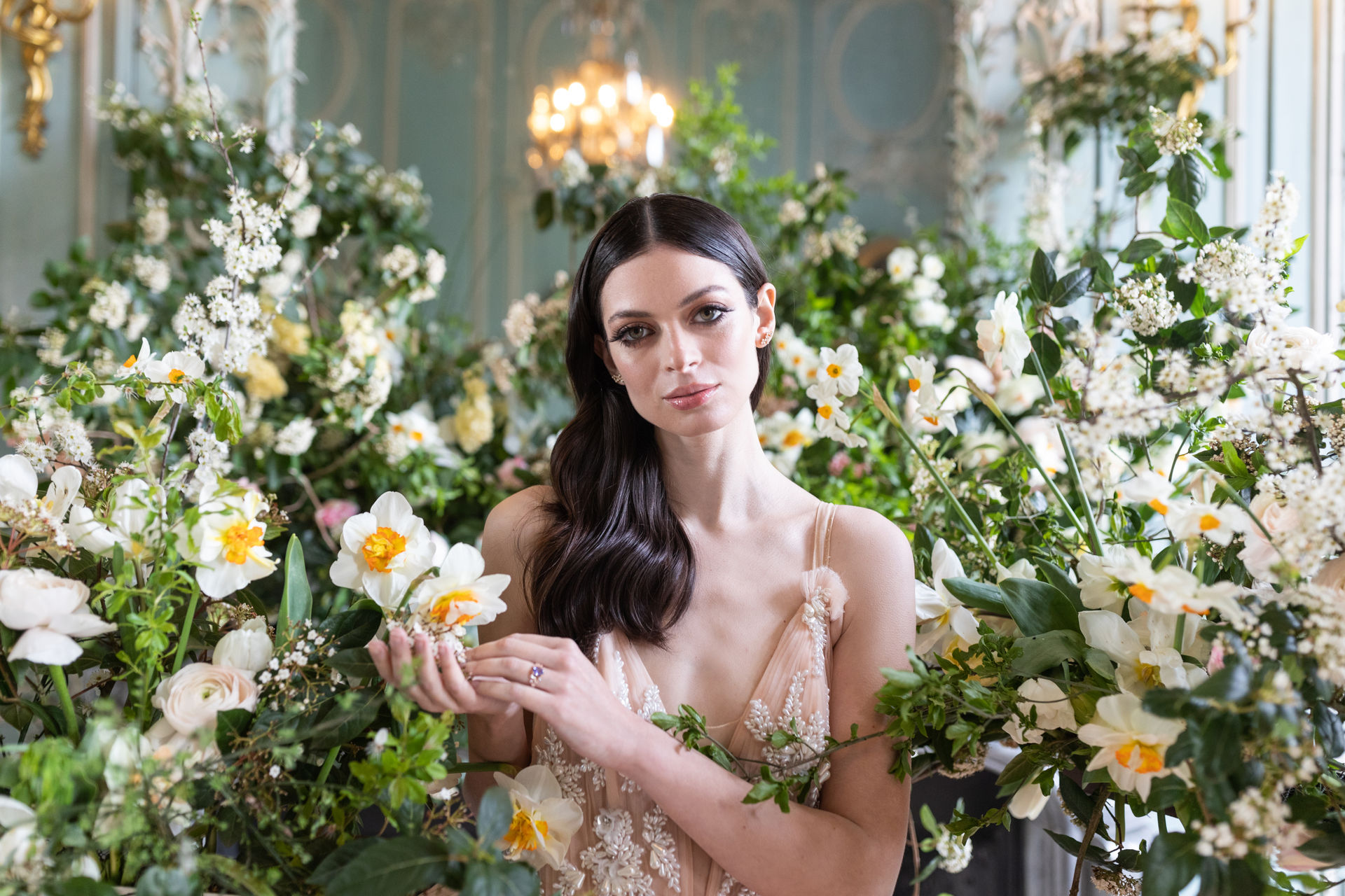 A close-up portrait of a bride surrounded by large-scale floral installations inside an ornate ballroom or salon with pale blue-grey panelled walls, gold gilded mouldings, and a gold chandelier visible in the background. The bride wears a blush/champagne spaghetti-strap gown with delicate floral lace or embroidery, and a coloured gemstone ring — likely amethyst or light purple — on her finger. Her dark hair is worn loosely waved over one shoulder, and she wears small ear cuff or stud jewellery. The floral arrangements surrounding her feature white and yellow-centred daffodils, white ranunculus, blush garden roses, white blossom branches, and abundant lush greenery, creating a garden-like installation effect in a classic French interior. The overall styling combines a romantic, garden-inspired floral palette of white, cream, soft yellow, and blush with the formal architectural backdrop of the venue.