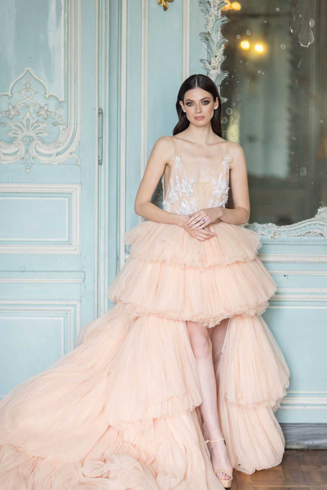 A bridal portrait of a single woman posed indoors against ornate pale blue-painted boiserie paneling and a gilded mirror, characteristic of a French chateau interior. She wears a blush peach tiered tulle ball gown with a high-low silhouette, thin spaghetti straps, a sheer bodice with silver crystal and beaded appliqué, and multiple voluminous ruffled tulle layers forming the skirt. Her dark hair is worn sleek and down, she has crystal stud earrings and a purple gemstone ring, and she wears nude strappy heeled sandals. The shot is a medium-length portrait with soft, diffused natural lighting complementing the warm peach tones of the gown against the muted blue backdrop.