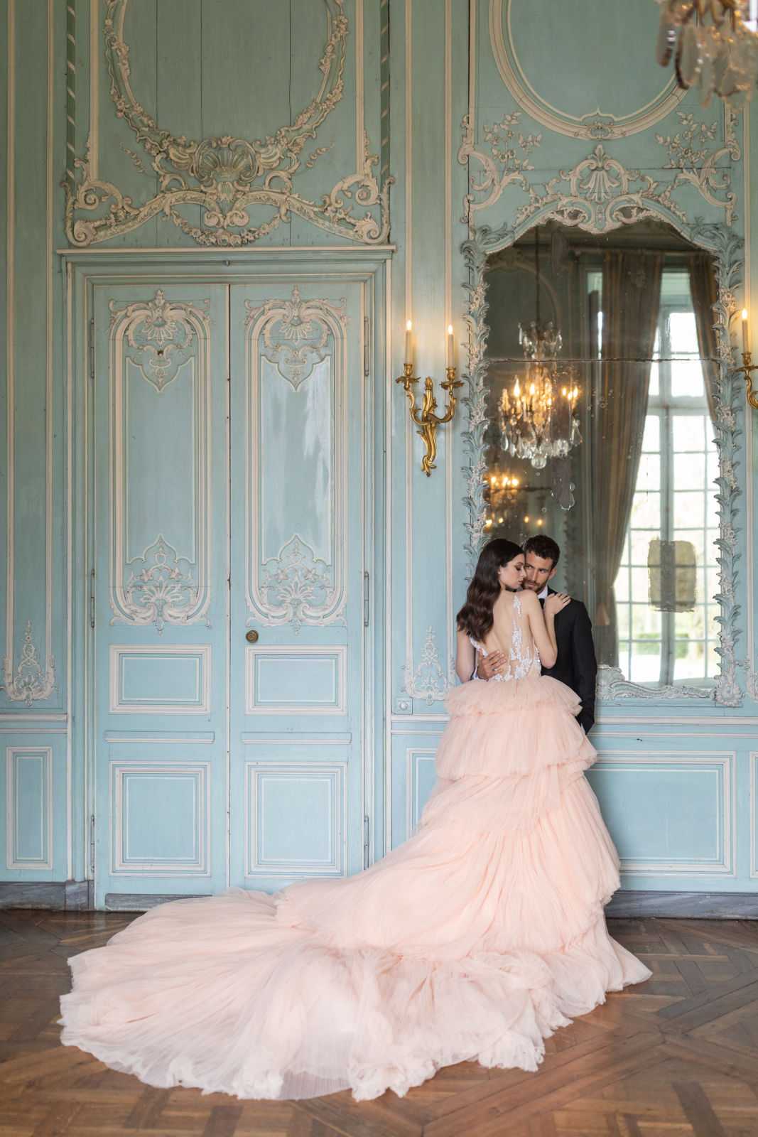 A couple poses intimately in a portrait shot inside a French chateau salon, with the bride leaning into the groom as he leans against the wall beside a large ornate mirror. The bride wears a blush pink ballgown with a lace illusion bodice and an extensively tiered, ruffled skirt with a long sweeping train spread across the herringbone parquet floor; the groom is dressed in a dark navy or black suit. The room features pale blue-green boiserie paneling with white and gold rococo plasterwork detailing, gold gilded wall sconces with lit candles, a large antique mirror with ornate carved framing, and a crystal chandelier reflected in the mirror. The overall styling is classic French baroque-inspired, and the wide portrait composition showcases both the full gown and the ornate architectural interior. Potential venue feature image.