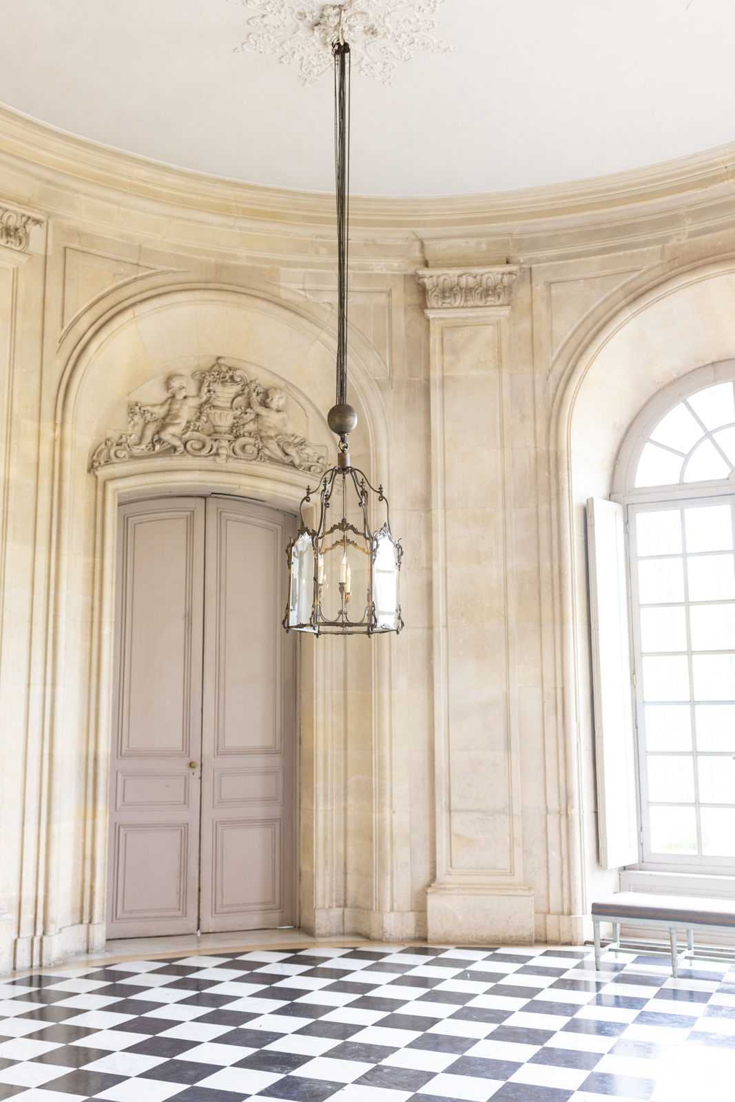 A venue interior shot with no people present, showing a grand classical French hall with cream-colored stone walls featuring carved bas-relief cherub ornamentation above an arched doorway with grey-painted double doors. A wrought-iron and glass lantern pendant light hangs from a white ornamental plaster ceiling medallion, fitted with candelabra-style bulbs. The floor is laid with a black and white diamond-pattern marble tile, and tall arched windows with white-painted frames allow natural light into the space. A simple modern bench is partially visible at the right edge of the frame. Wide shot composition. Potential venue feature image.