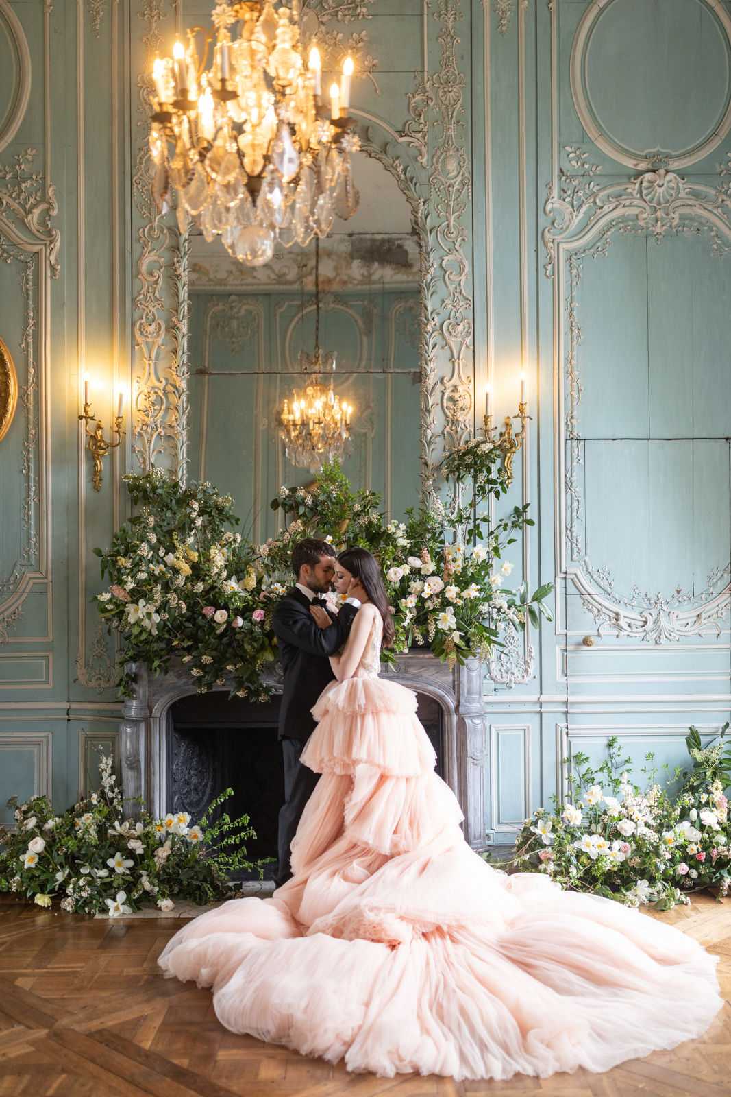 A couple shares an intimate forehead-to-forehead moment in a portrait shot taken indoors in a French château-style ballroom with powder blue painted boiserie paneling, ornate gilt plasterwork moldings, and a carved marble fireplace. The bride wears a voluminous blush pink tiered tulle ballgown with an extensive floor-sweeping train, while the groom is dressed in a classic black tuxedo with bow tie. The fireplace mantel and surrounding floor are dressed with lush, overflowing arrangements of white and cream roses, blush ranunculus, white narcissus, wisteria, and abundant green foliage, creating a garden-like installation that extends across the parquet herringbone floor. A large crystal chandelier hangs overhead and gilt candle wall sconces flank a floor-to-ceiling mirror above the fireplace, reflecting the chandelier and adding warm candlelight tones to the room's classic French interior styling. Potential venue feature image.