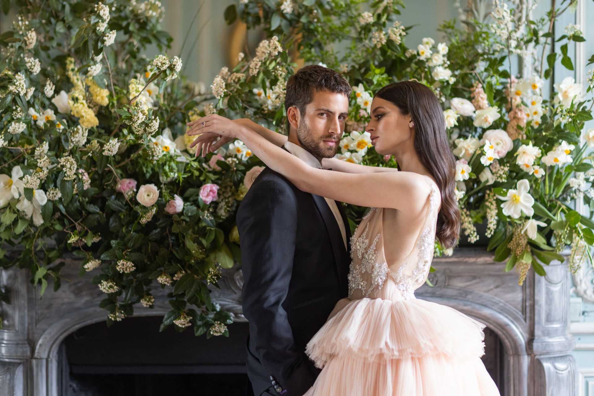 A couple poses indoors in front of an ornate marble fireplace that has been dressed with a large, lush floral installation. The bride wears a blush pink tiered tulle ballgown with a beaded, low-back bodice, and her arm is draped around the groom's shoulder as the two face each other closely. The groom is dressed in a black tuxedo jacket with a champagne lapel. The floral arrangement covering the fireplace mantel includes white daffodils, blush and white ranunculus, soft pink garden roses, yellow blossom clusters, white Queen Anne's lace-style blooms, and abundant greenery. The room features pale sage-painted walls with classical architectural detailing visible at the edges of the frame. This is a medium portrait-style shot with the couple centered against the floral backdrop.