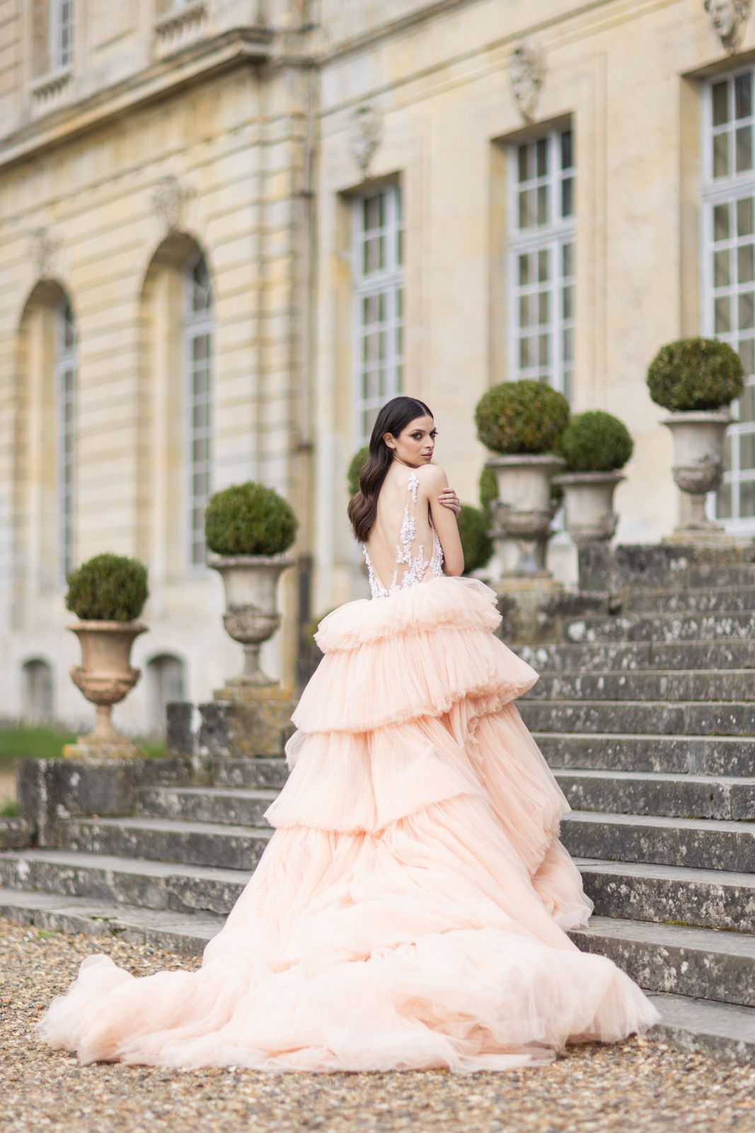A bridal portrait shot outdoors on the stone steps of a French chateau, featuring a bride in a voluminous blush pink tiered tulle ballgown with a sheer lace-appliqué open back and an extended train that spreads across the gravel. The bride, with dark wavy hair worn down, glances back over her shoulder toward the camera. The chateau facade features classical French architecture with tall windows, and the steps are flanked by trimmed topiary shrubs in large stone urns. The image is a full-length portrait with a shallow depth of field that keeps the bride sharp against the softly blurred architectural background. Potential venue feature image.