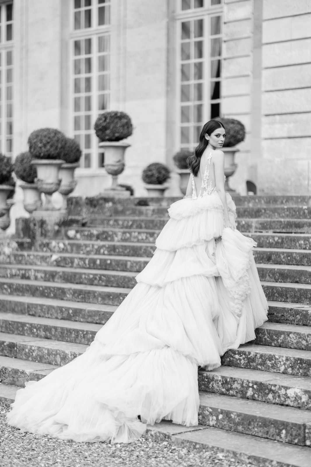 This is a black-and-white bridal portrait of a single bride standing on broad stone steps in front of a classical French chateau facade. She is positioned three-quarters away from the camera, glancing back over her shoulder, with her dark hair styled in loose waves. Her gown features a deeply open back with lace or embroidered bodice detailing and a dramatically voluminous tiered tulle ballgown skirt with a long cathedral-style train that cascades down multiple steps. The image shows strong contrast between the bright light tones of the dress and the mid-grey tones of the stone steps and building. Large ornamental stone urns with clipped topiary are visible along the terrace behind her. The composition is a full-length portrait shot with a shallow depth of field, keeping the bride sharp while the chateau architecture recedes softly in the background. Potential venue feature image.