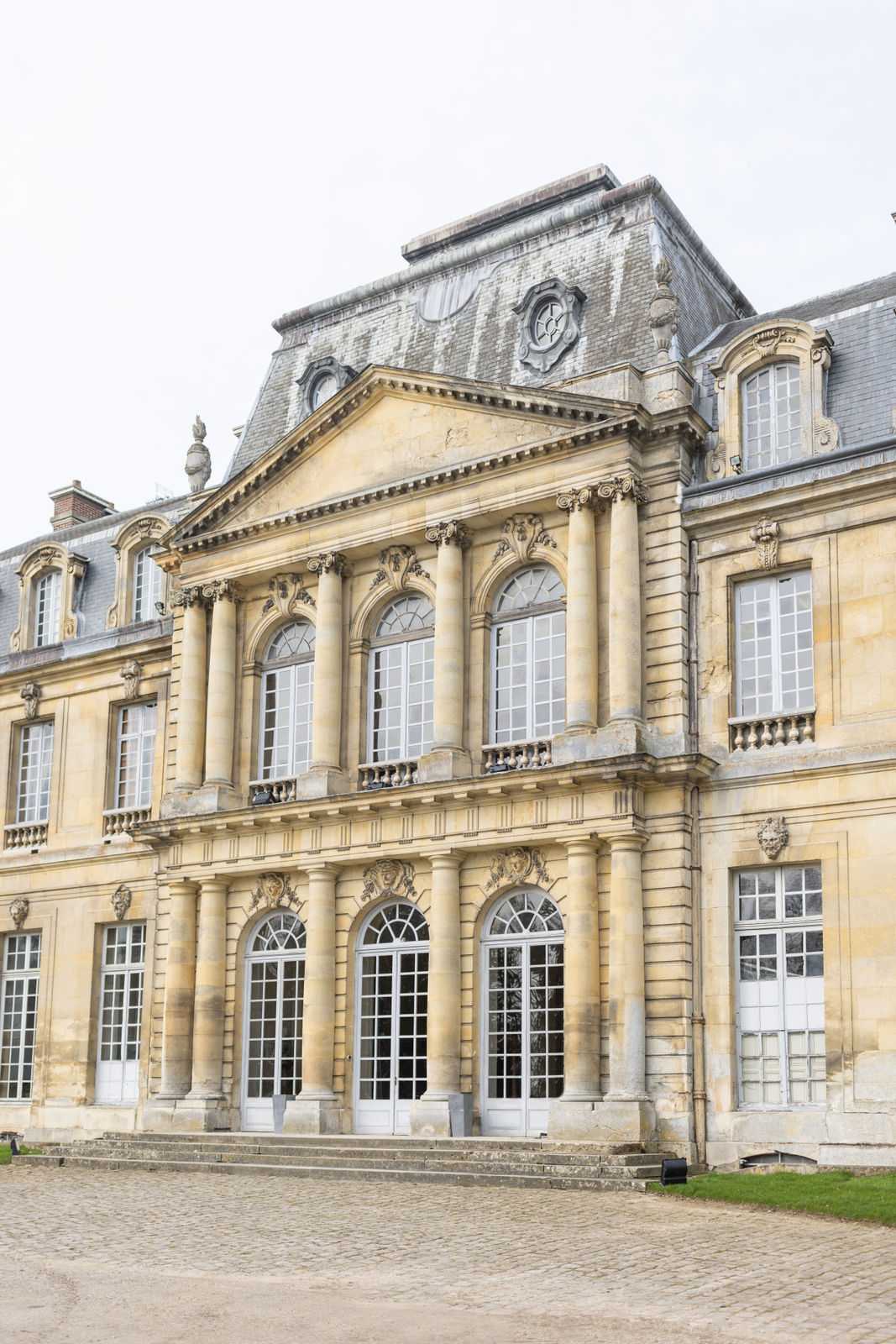 Wide shot of the exterior facade of a large French classical chateau, featuring warm golden limestone construction with two stories of arched windows framed by decorative columns with carved keystones, a central triangular pediment above the upper level, and a grey mansard roof with ornate stone detailing. The building includes tall arched ground-floor doors with white painted frames, a cobblestone courtyard in the foreground, and stone steps leading to the main entrance. No people or wedding elements are visible in this image. Potential venue feature image.