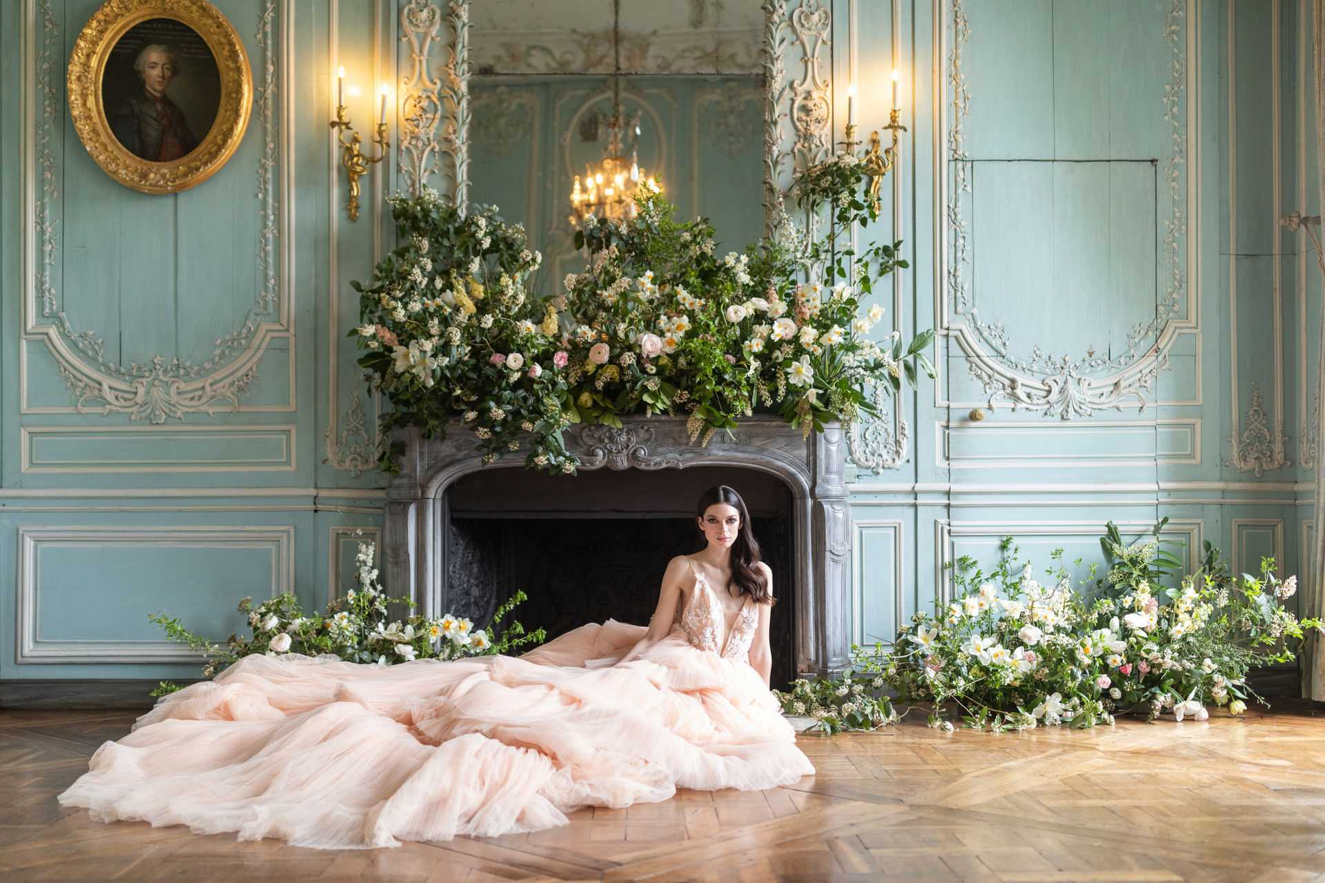 A bridal portrait shoot set inside a French château salon, featuring a bride seated on a parquet herringbone floor in front of an ornate carved stone fireplace. The bride wears a blush pink ballgown with a deep V-neck lace bodice and a voluminous tulle skirt spread across the floor. The fireplace mantel is heavily decorated with an overflowing floral arrangement of white ranunculus, cream and blush roses, white narcissus, and abundant greenery, with additional ground-level floral clusters flanking the fireplace on both sides. The room features pale duck-egg blue painted boiserie wall paneling with white plaster molding details, gilt brass wall sconces with lit candles, a crystal chandelier visible in the fireplace mirror reflection, and a gilded oval portrait painting on the left wall. The styling theme is French classical with a garden-inspired, organic floral approach. Wide portrait shot centered on the bride against the fireplace backdrop. Potential venue feature image.