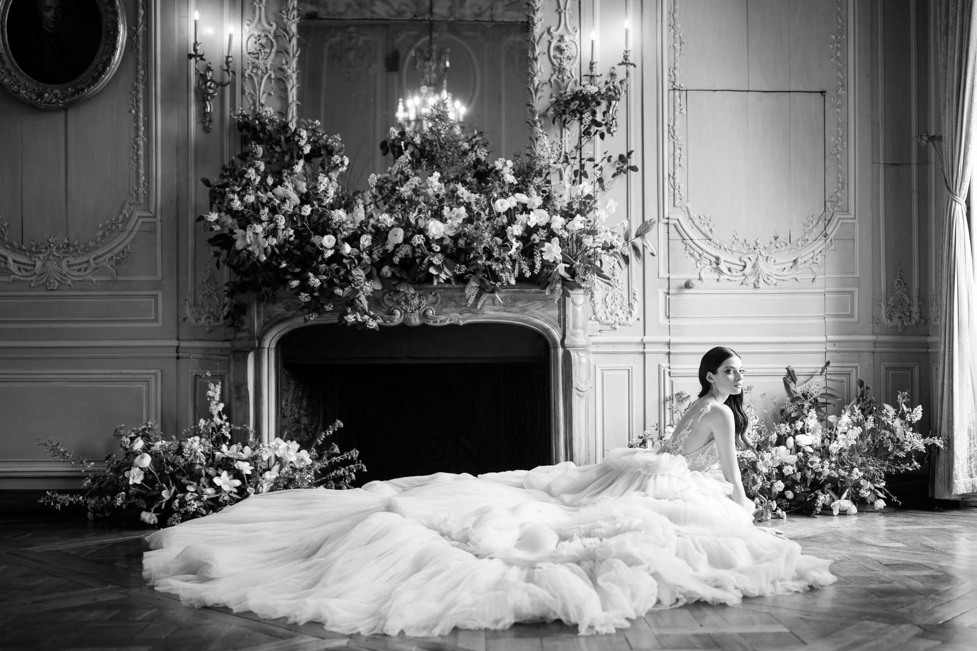 This black-and-white bridal portrait shows a bride seated on a parquet floor in front of an ornate carved fireplace inside what appears to be a French chateau ballroom or grand salon. She wears a voluminous tulle ball gown with a lace bodice and an extremely long, ruffled train that spreads widely across the floor in front of her; she looks over her shoulder with her dark hair worn down. The fireplace mantel is densely covered in an oversized floral installation featuring light-toned blooms — identifiable as ranunculus, roses, and trailing greenery — and additional loose floral clusters are arranged on the floor to both sides of the fireplace. The room features heavily carved boiserie wall paneling, gilded wall sconces with lit candles, an oval portrait painting, and a crystal chandelier reflected in a mirror above the mantel, all rendered in high-contrast tones. The composition is a wide environmental portrait that places the bride as a secondary element to the full architectural and floral scene. Potential venue feature image.