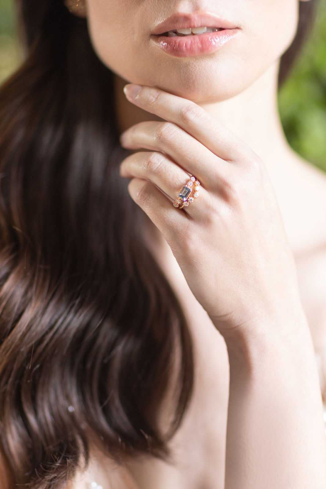 A close-up detail portrait of a bride's hand resting against her chin and neck, showcasing a stacked ring set on her ring finger. The rings feature a rose gold setting with a rectangular emerald-cut grey/blue center stone, accompanied by smaller accent stones including what appear to be pink and white diamonds or gemstones, styled as a bridal ring stack. The bride has long dark brown wavy hair and a bare shoulder visible, suggesting a strapless or low-cut dress. The background is softly blurred with hints of greenery. The composition is a tight portrait-style detail shot focused on the jewelry.