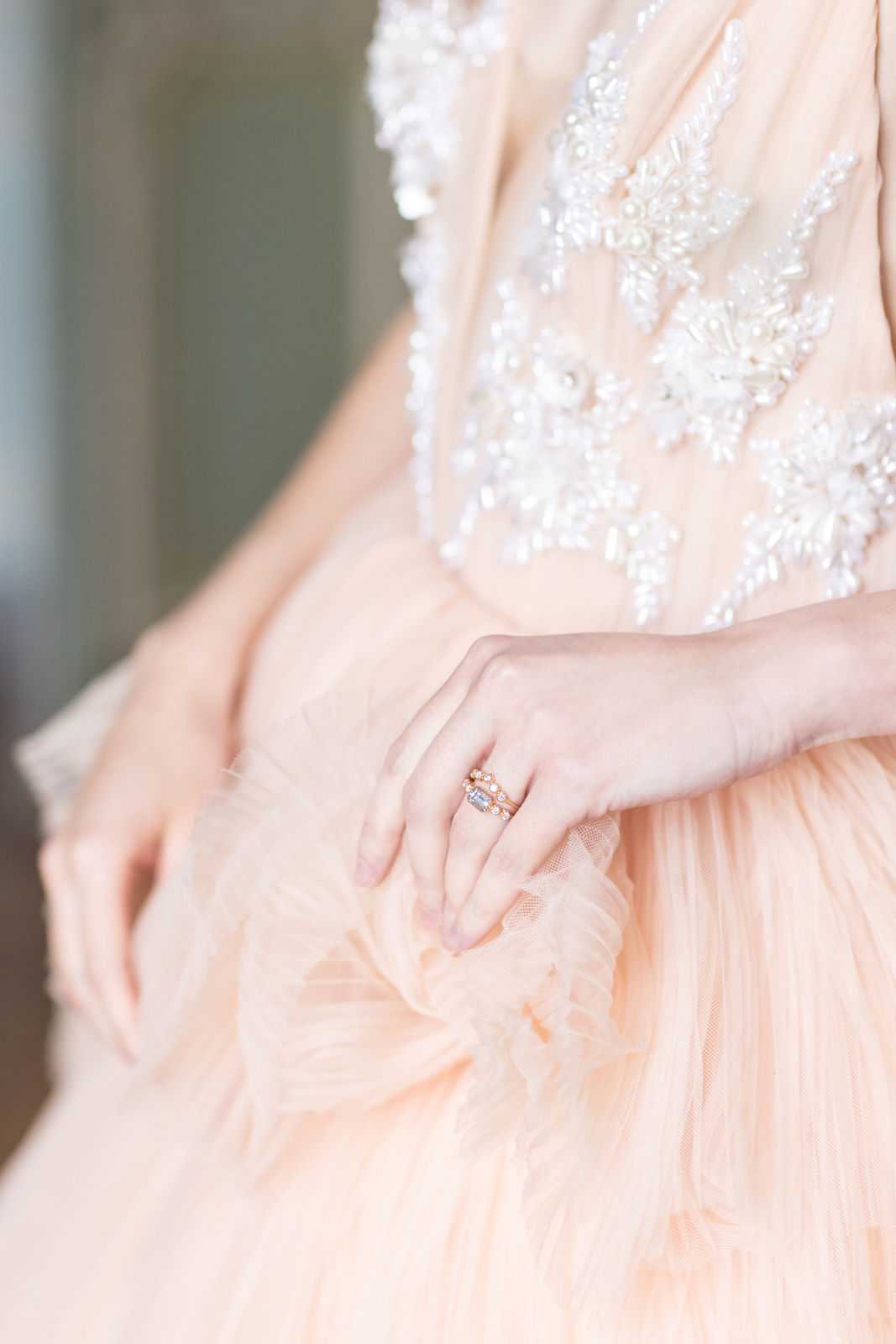 A close-up detail shot of a bride's hands resting on the skirt of a blush peach tulle wedding dress with a heavily beaded and pearl-embellished bodice featuring floral and leaf motifs in white and ivory. The bride wears a stacked ring set on her left ring finger, consisting of a rose gold engagement ring with an emerald-cut center stone accompanied by a diamond-set band. The layered tulle skirt has a soft ruffled texture in the same blush peach tone as the bodice. The background is softly blurred, keeping the focus entirely on the dress detail and rings.