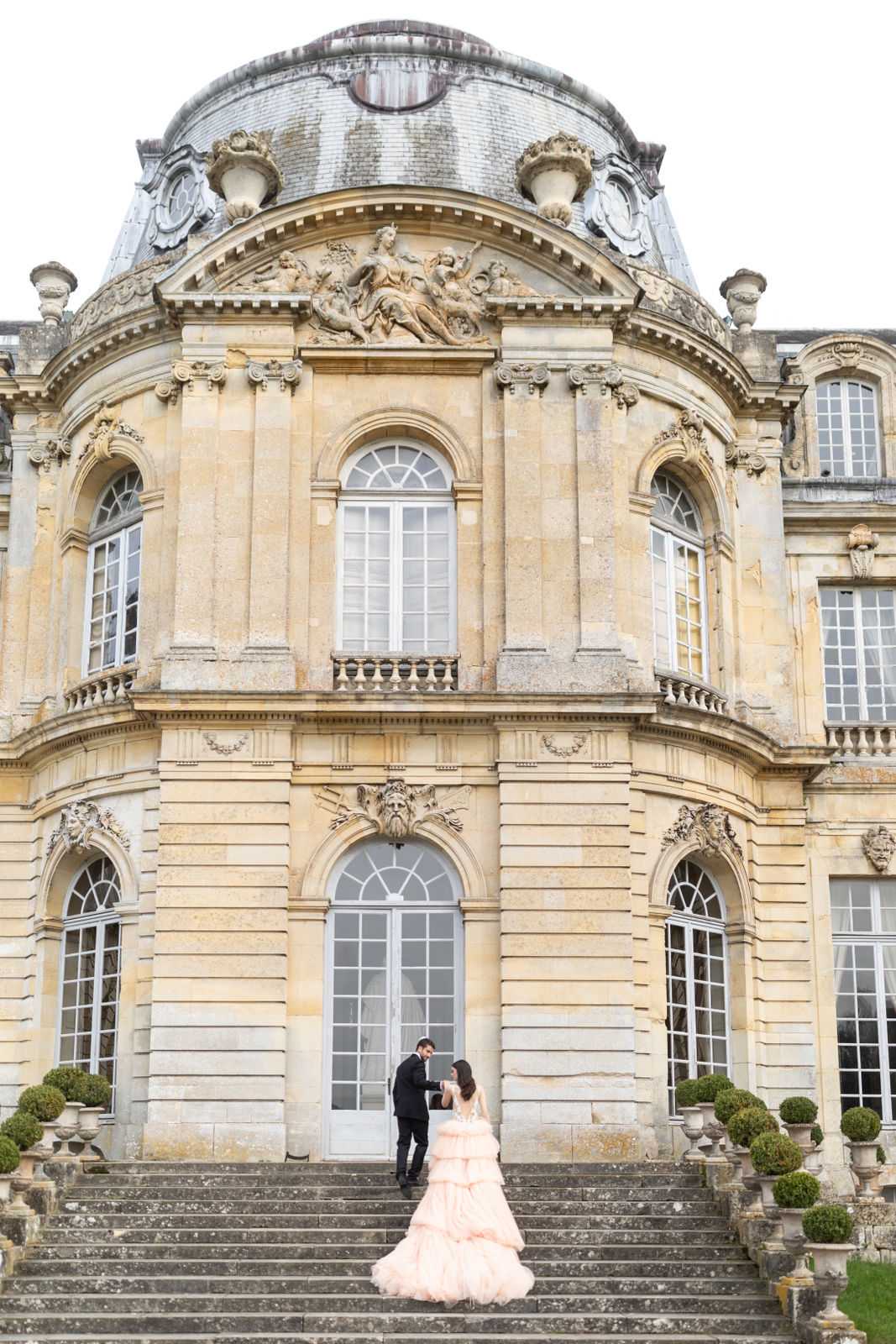 A couple poses on the grand exterior staircase of a French classical chateau during a portrait session. The groom, dressed in a black tuxedo, takes the bride's hand as they stand on the steps facing the building's entrance. The bride wears a voluminous blush pink tiered ballgown with a long train that cascades down the stone steps. The chateau facade is ornate French Baroque architecture featuring carved stone relief sculptures, arched windows, a domed roof, and decorative balustrades, with small trimmed topiary urns flanking the staircase. The composition is a wide portrait shot that emphasizes the scale of the building against the small figures of the couple. Potential venue feature image.