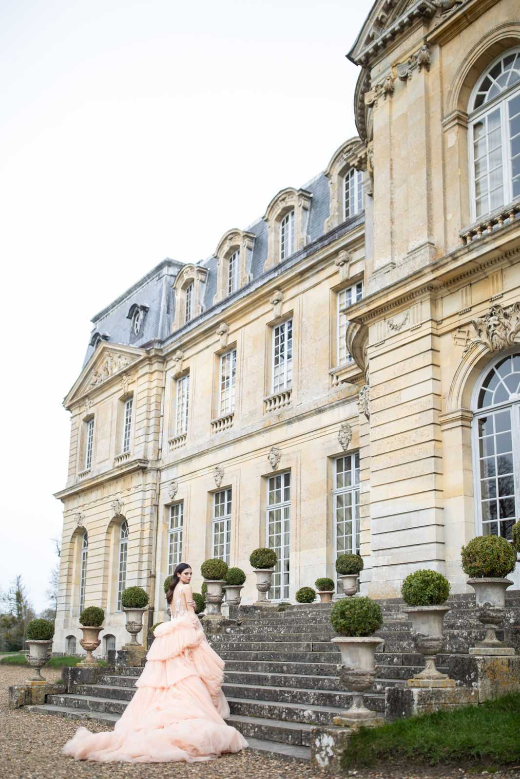 A bride poses alone on the gravel grounds at the base of the stone entrance steps of a grand French classical chateau, shot from a wide-angle perspective that emphasizes the full scale of the building's facade. She wears a voluminous blush pink tiered ballgown with a dramatically long ruffled train spread across the ground, and a fitted embellished bodice; she is turned slightly away from the camera. The chateau features ornate carved stone detailing, tall French windows, a mansard slate roof with dormer windows, and stone urns holding clipped topiary box balls lining the entrance steps. Potential venue feature image.