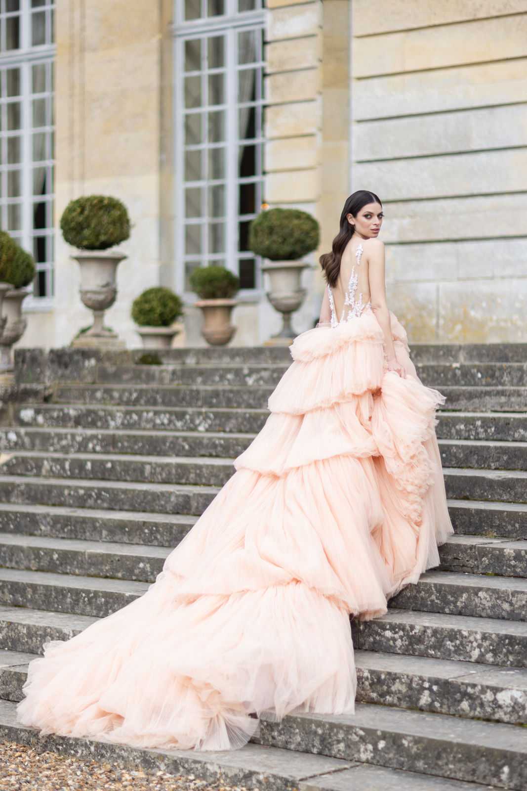 A bride poses on the stone exterior staircase of a French chateau, looking back over her shoulder toward the camera. She wears a voluminous blush pink tulle ball gown with multiple tiered ruffled layers and a sheer, lace-embellished low back, with a long dramatic train cascading down the steps. The chateau facade features tall French windows and large stone urns with trimmed topiary boxwood balls. The shot is a full-length portrait taken from behind, emphasizing the scale and layered construction of the gown against the classical architecture.