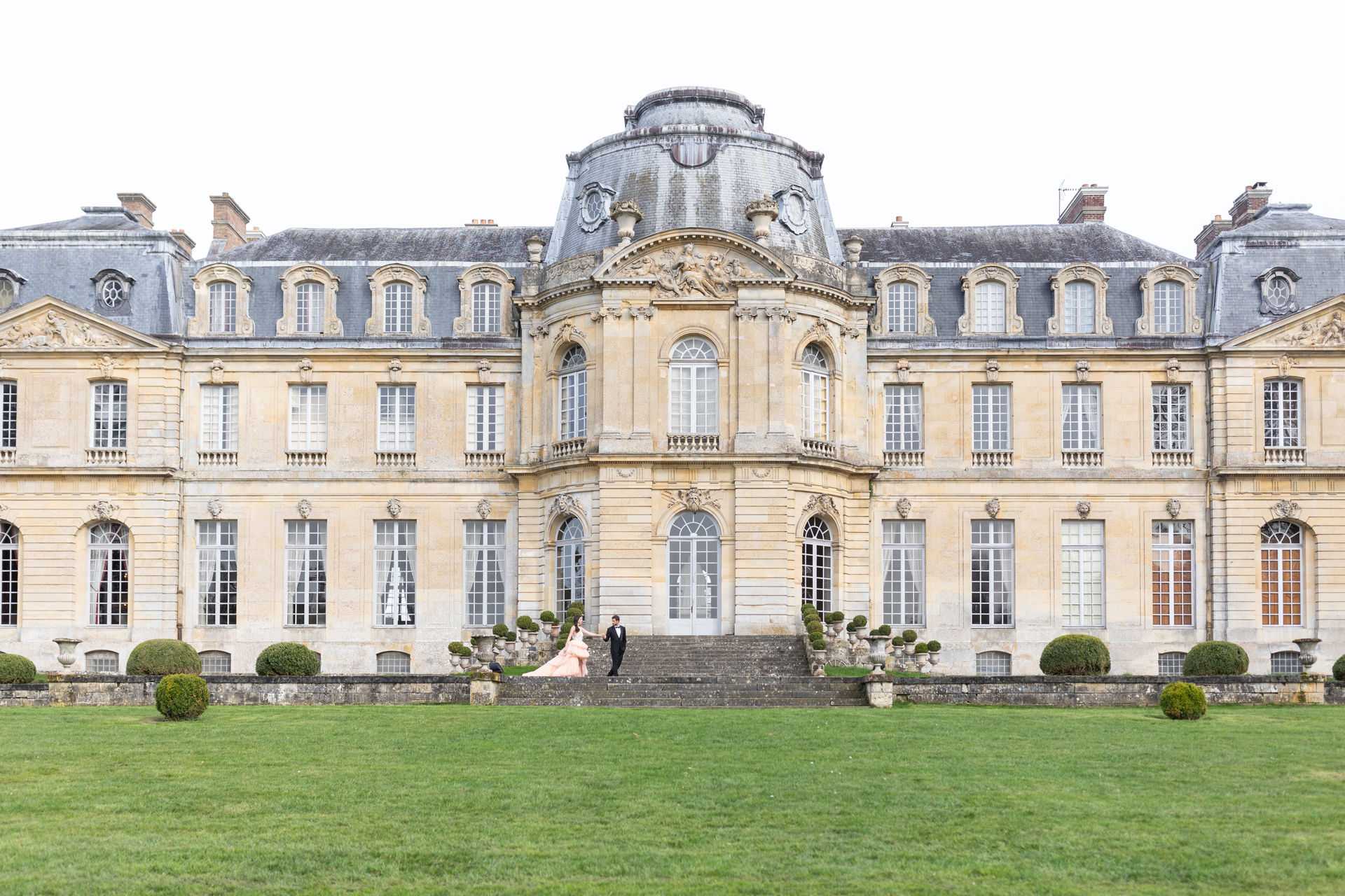 A couple poses on the stone steps of a large French classical chateau during what appears to be a wedding portrait session. The bride wears a full-length blush pink ball gown and the groom is dressed in a black tuxedo; they are holding hands on the central staircase of the building. The chateau facade is a wide, symmetrical structure in pale golden limestone with a mansard roof, ornate carved stonework, and a projecting central pavilion topped with a dome — consistent with 17th or 18th century French architecture. Formally clipped topiary shrubs and stone urns line the terrace balustrade. The composition is a wide establishing shot taken from across the lawn, making the couple appear small in scale against the grand facade. Potential venue feature image.
