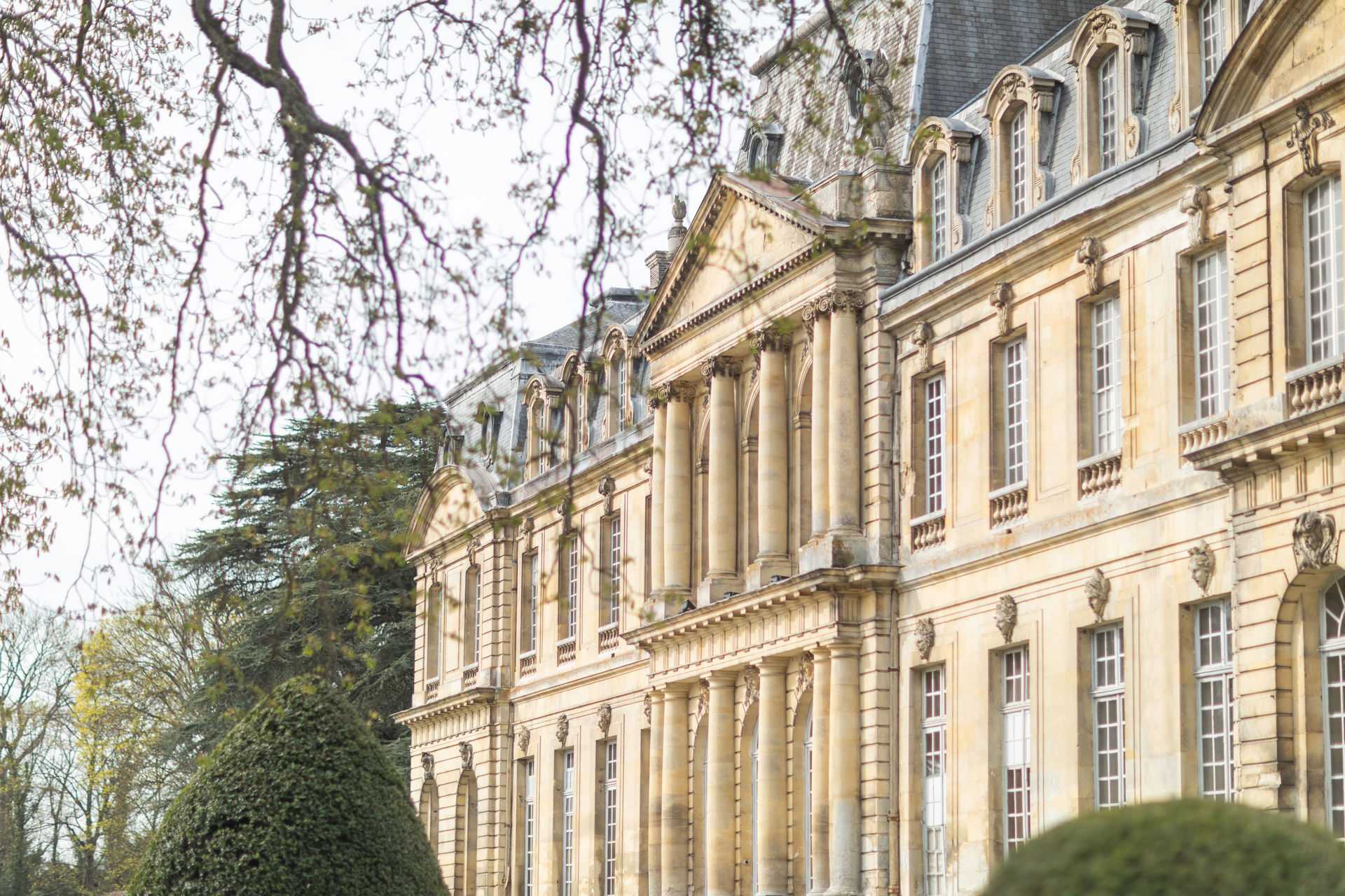 An exterior architectural shot of a grand French chateau, photographed from a slightly angled perspective that emphasizes the facade's classical detailing. The building features warm honey-toned limestone construction with tall pilasters, ornate carved stone medallions, a central pediment with columns, large arched and rectangular windows, and a slate mansard roof with dormer windows. No people are present in the frame; the image focuses entirely on the architecture and grounds. The composition is a wide exterior shot with bare-branched trees framing the upper left portion of the image, and manicured topiary visible at the lower right. Potential venue feature image.