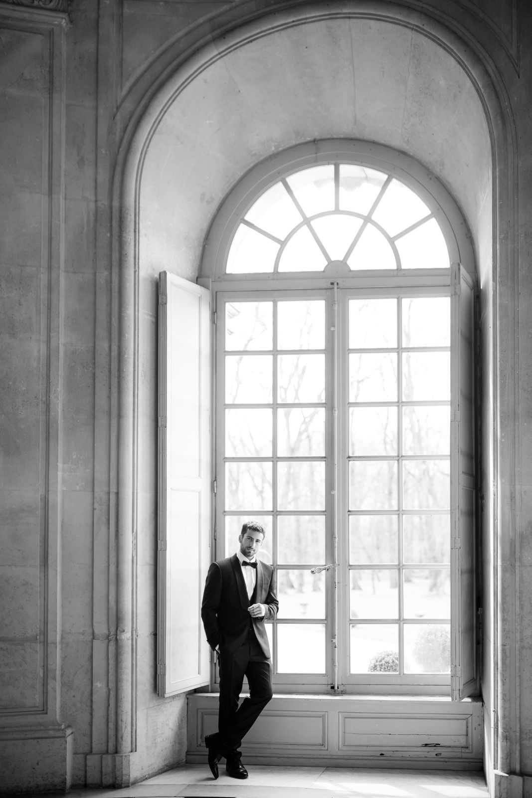 A black-and-white full-length portrait of a groom standing in front of a tall arched window inside a grand classical interior, likely a French chateau or palace. The groom is dressed in a dark tuxedo with a bow tie and dress shoes, leaning casually against the open window frame with one hand in his pocket. The window features fanlight glazing at the top and multi-pane casement doors, flooding the space with bright natural light that creates strong tonal contrast against the carved stone moldings of the arched recess. The composition is a wide vertical portrait, with the groom positioned in the lower-center third of the frame, emphasizing the architectural scale of the setting.