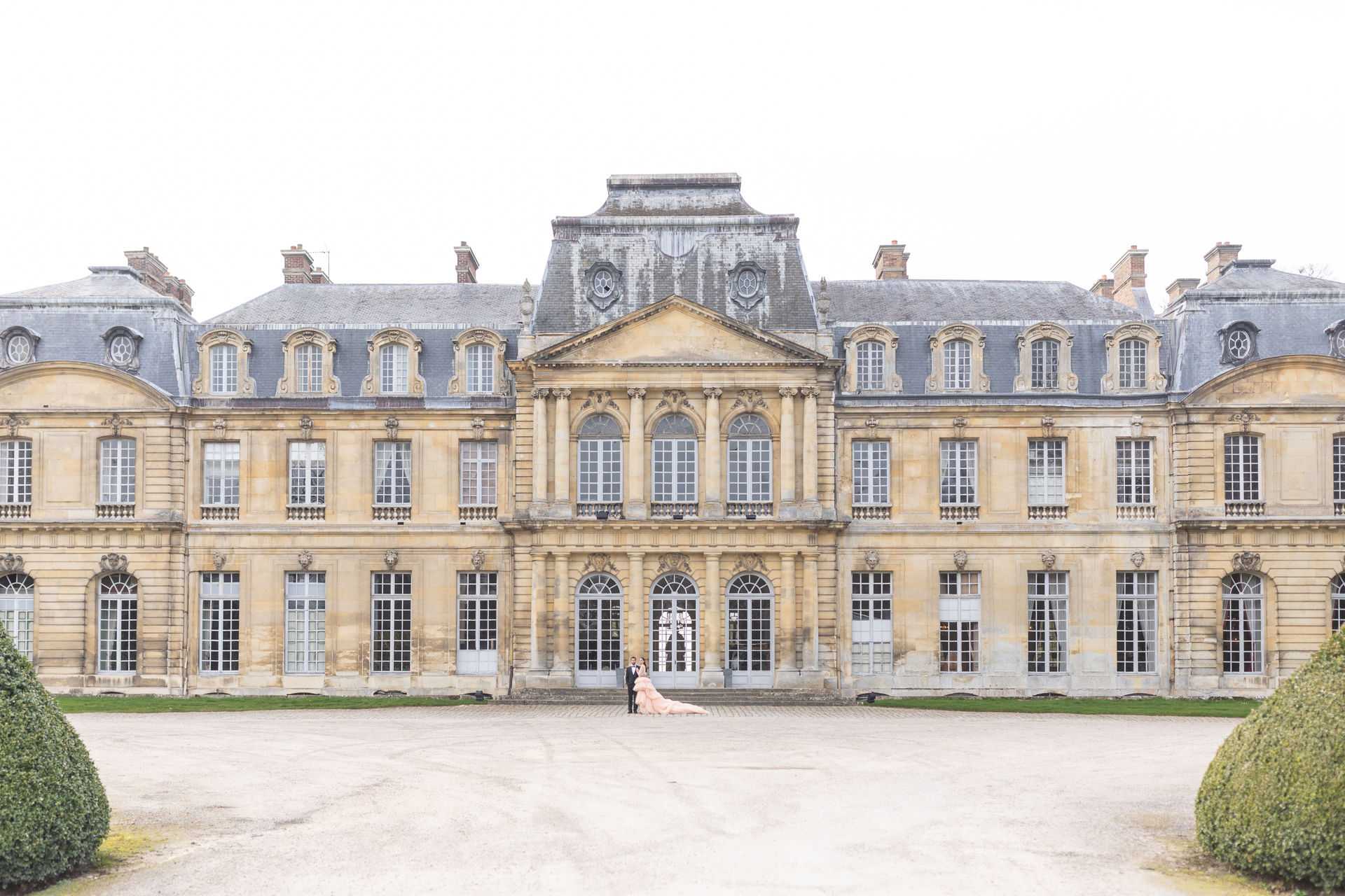 A wide-shot couple portrait taken outdoors in front of a grand French classical chateau with warm golden limestone facades, mansard slate roofs, and symmetrical baroque-style architecture across three stories. The couple stands at the central entrance steps of the chateau, appearing very small in scale against the expansive building, emphasizing the scale of the venue. The bride wears a voluminous blush pink ball gown with an extremely long train that spreads dramatically across the gravel forecourt, while the groom is dressed in a dark suit. Manicured boxwood topiaries frame the foreground, and the overall composition is centered and symmetrical, shot from across the gravel courtyard. Potential venue feature image.