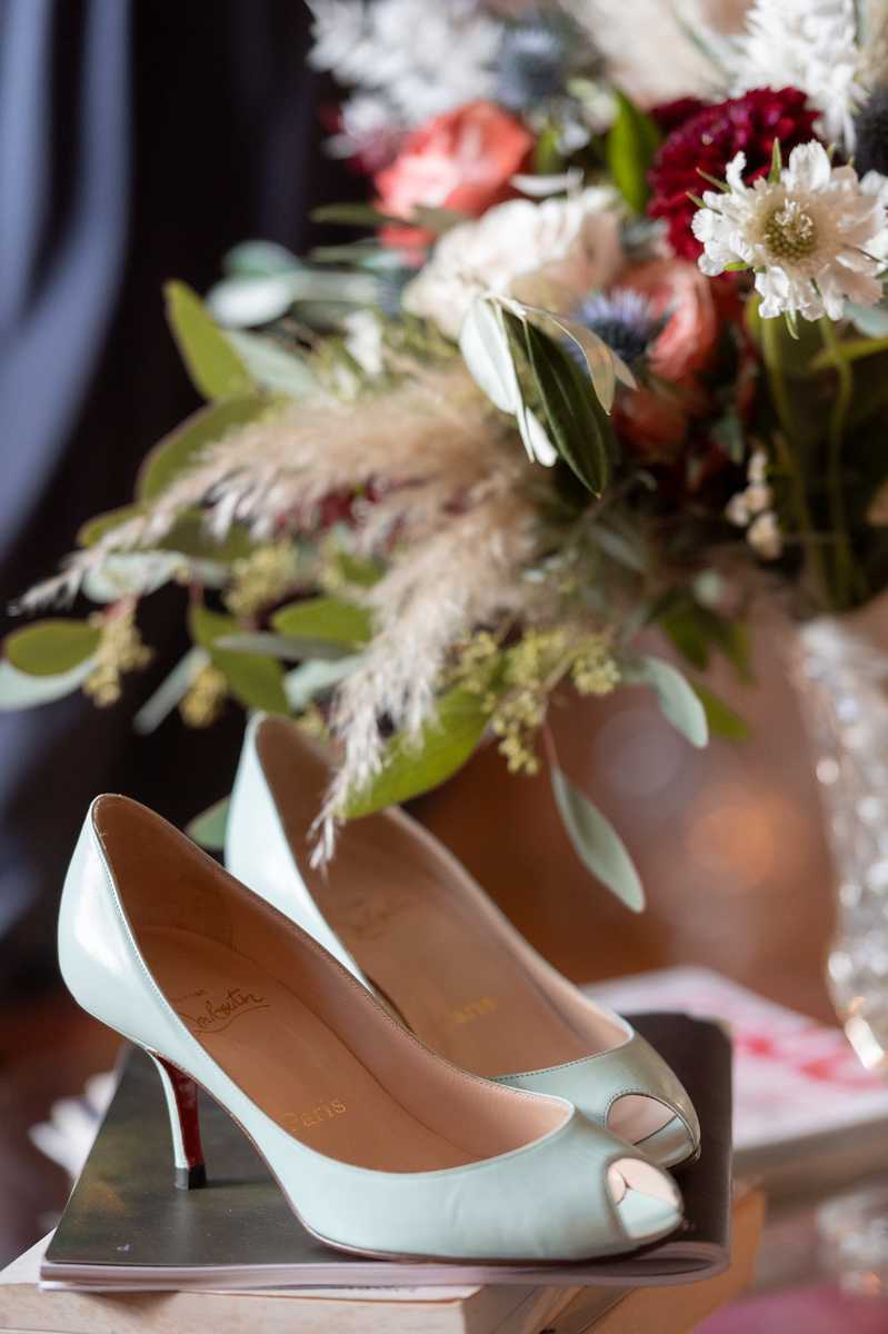Close-up detail shot of a pair of Christian Louboutin peep-toe heels in pale mint green with the signature red sole, placed on a small tray or book. In the soft-focus background sits a bridal bouquet featuring deep burgundy carnations or ranunculus, coral blooms, white scabiosa, pampas grass, eucalyptus, and mixed greenery, arranged in a boho-inspired, textured style. The overall color palette mixes mint, burgundy, coral, and ivory, suggesting a bold, eclectic wedding aesthetic.
