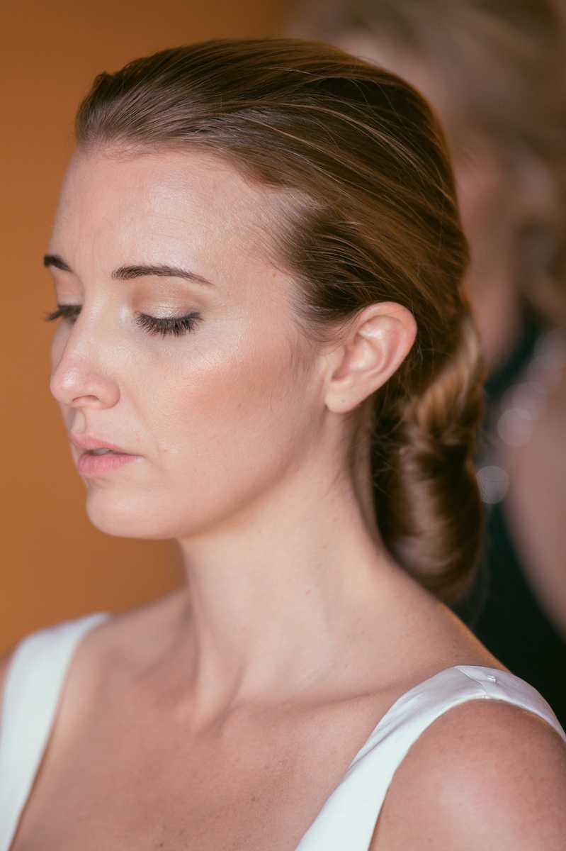A close-up portrait of a bride during the getting-ready phase, captured in profile with her eyes closed. She wears a sleek, slicked-back low chignon hairstyle and minimal makeup with defined brows, a subtle shimmer eye, and a nude lip. The top strap of a white dress is visible at the bottom of the frame. A second person, likely a hair or makeup stylist, is visible but out of focus in the background against a warm amber-toned wall.