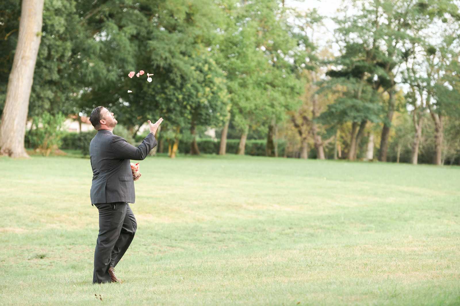 A man in a dark charcoal suit with brown dress shoes stands alone on a large open lawn, tossing pink and white flower petals into the air above his head while holding what appears to be a basket or container of petals in his other hand. He is looking upward with his face tilted toward the falling petals. The shot is taken outdoors in a wide-angle portrait style, capturing his full figure against a backdrop of mature trees and manicured hedging. The scene appears to be a candid or playful moment, likely during a cocktail hour or pre-ceremony setup on a chateau or estate property.