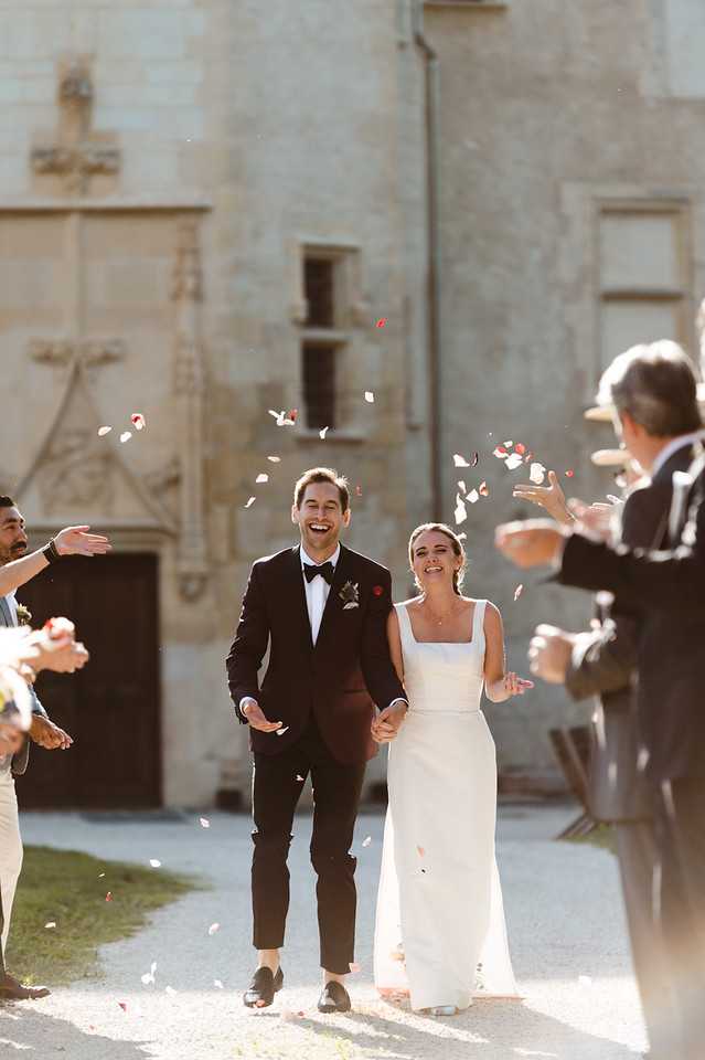 The bride and groom walk hand-in-hand through a confetti exit as guests on either side toss white and red rose petals over them. The groom wears a navy tuxedo with black bow tie and a red boutonniere, while the bride wears a sleek ivory slip-style gown with a square neckline. The setting is outdoor in front of a historic stone building with a large wooden door and carved stonework, consistent with a French chateau or chapel exterior. The shot is a medium portrait framing the couple in the center with blurred guests on both sides, captured in bright natural daylight.