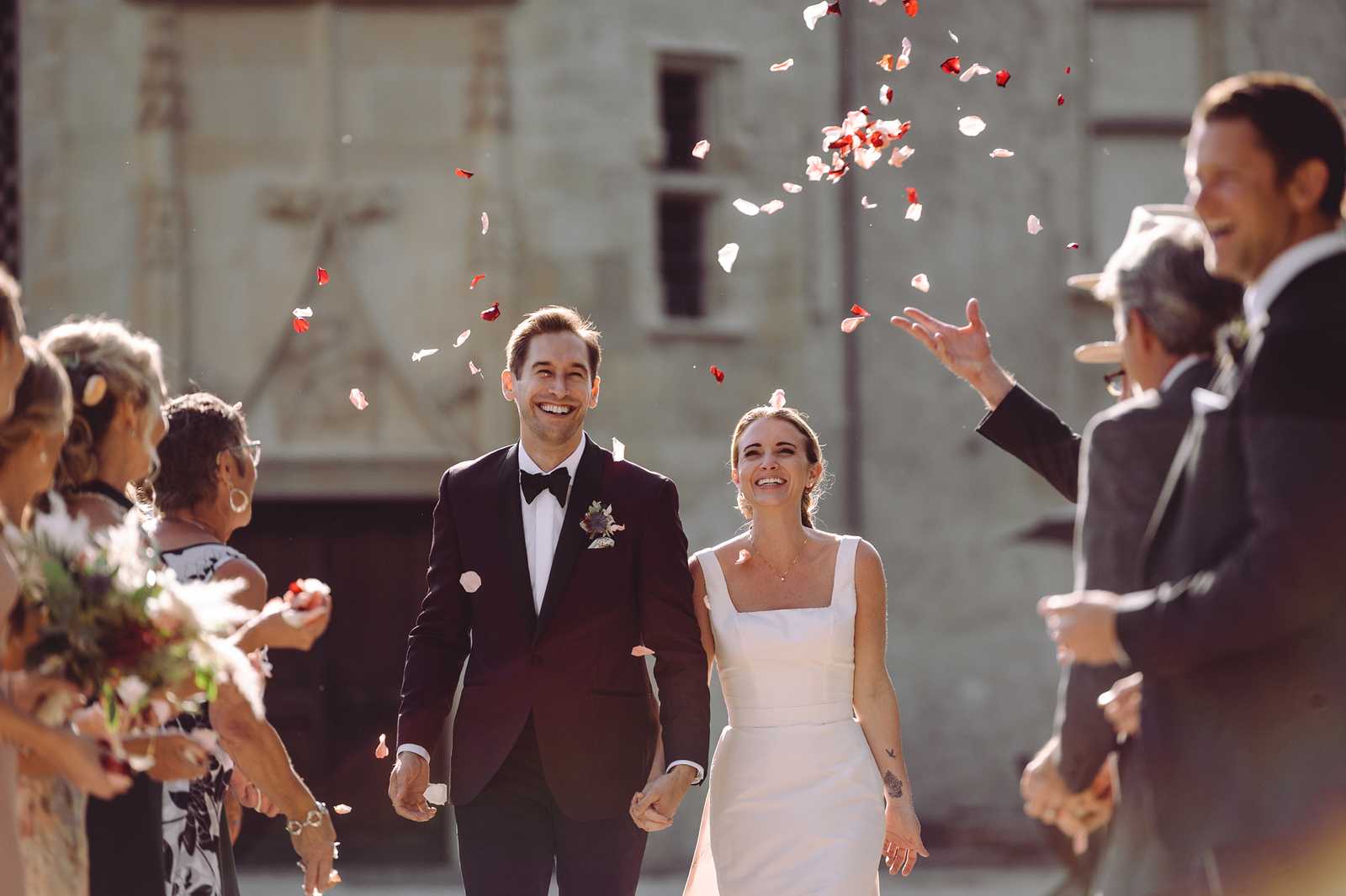 The couple is walking through a petal toss exit following their ceremony, outdoors in front of a large stone or rendered building with aged walls. Guests line both sides of the path, throwing a mix of red and blush pink rose petals into the air. The groom wears a deep navy or black tuxedo with a black bow tie and a small floral boutonniere, and the bride wears a minimalist ivory square-neck fitted dress with thick straps. Both are holding hands and laughing openly. Approximately six to eight guests are visible on either side, dressed in suits and mixed attire. The shot is a medium portrait-style image taken at ground level with the background softly out of focus, captured in warm natural backlight.