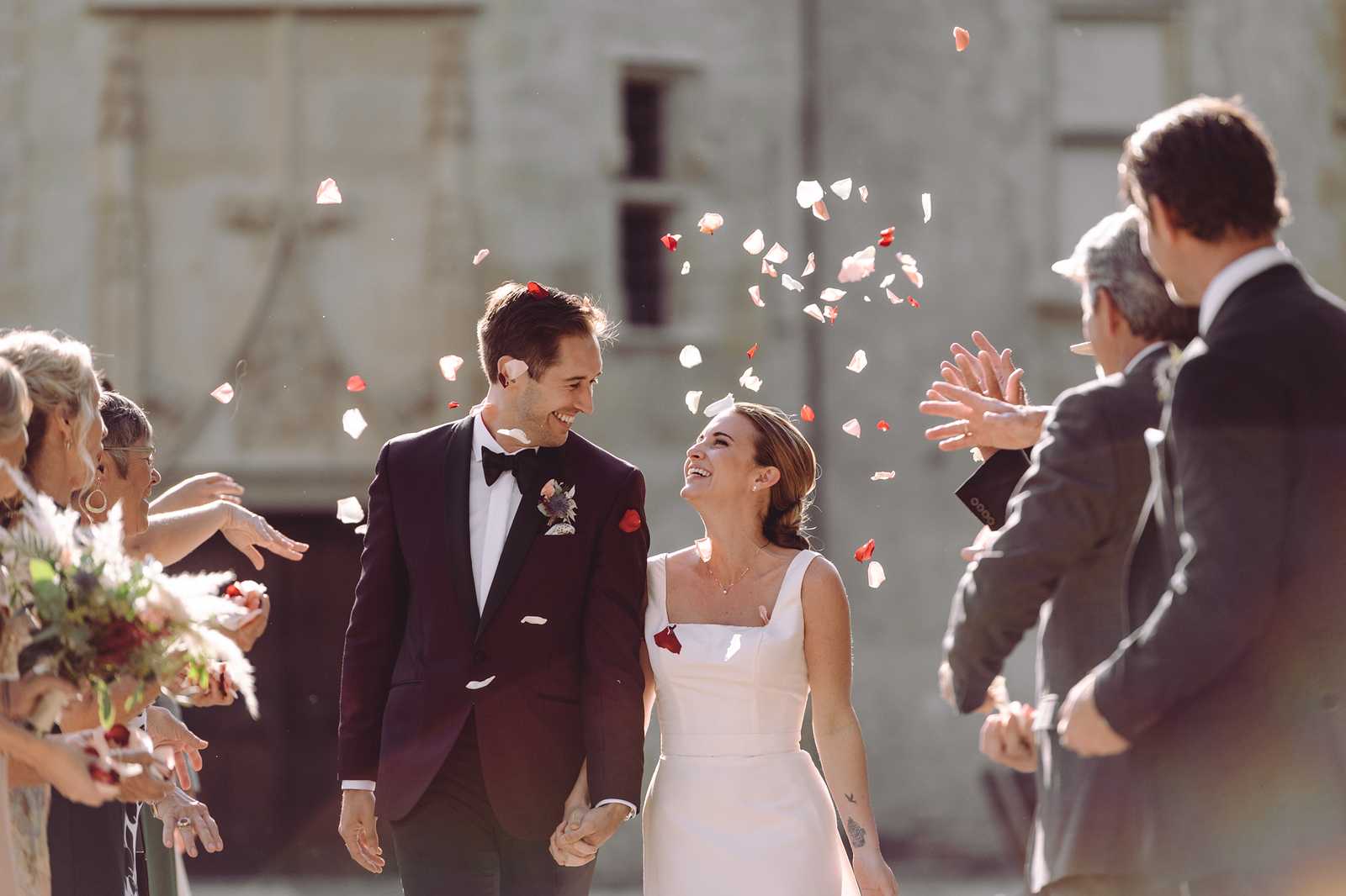 A couple walks through a petal toss exit outdoors, surrounded by approximately six to eight guests on either side throwing red and blush rose petals into the air. The groom wears a deep burgundy tuxedo jacket with black lapels, a black bow tie, white dress shirt, and a boutonniere featuring dried florals with a red accent; the bride wears a sleeveless ivory dress with a square neckline and a clean, minimal silhouette. The guests are dressed in dark formal attire, and a bridesmaid on the left holds a bouquet featuring pampas grass, deep red blooms, and blush tones. The shot is a medium portrait taken at eye level with a shallow depth of field, keeping the couple sharp while the background building and guests are softly blurred in warm natural backlight.