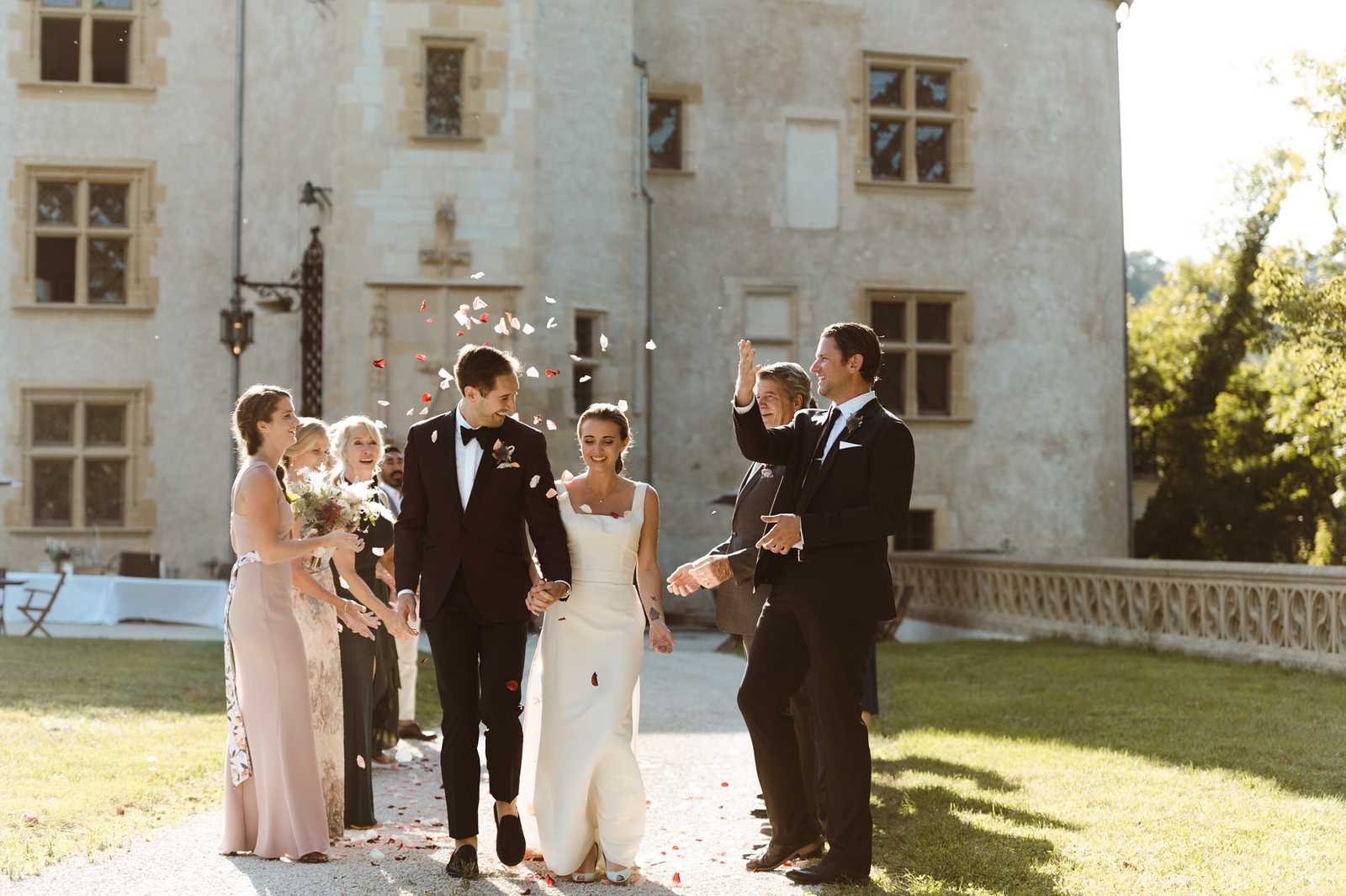 The bride and groom walk together along a gravel path in a post-ceremony recessional outside a French chateau, while approximately six guests line both sides throwing white and red rose petals over the couple. The bride wears a sleek, fitted ivory gown with a square neckline and a small train, while the groom is dressed in a black tuxedo with a bow tie. Guests on the left include a bridesmaid in a blush pink floor-length dress holding a bouquet with burgundy and neutral tones, alongside other guests in formal attire; guests on the right, in dark suits, are actively tossing petals. The scene is shot outdoors in warm late-afternoon sunlight against the facade of a stone chateau with arched windows and a decorative balustrade terrace visible in the background, captured in a wide candid portrait style.