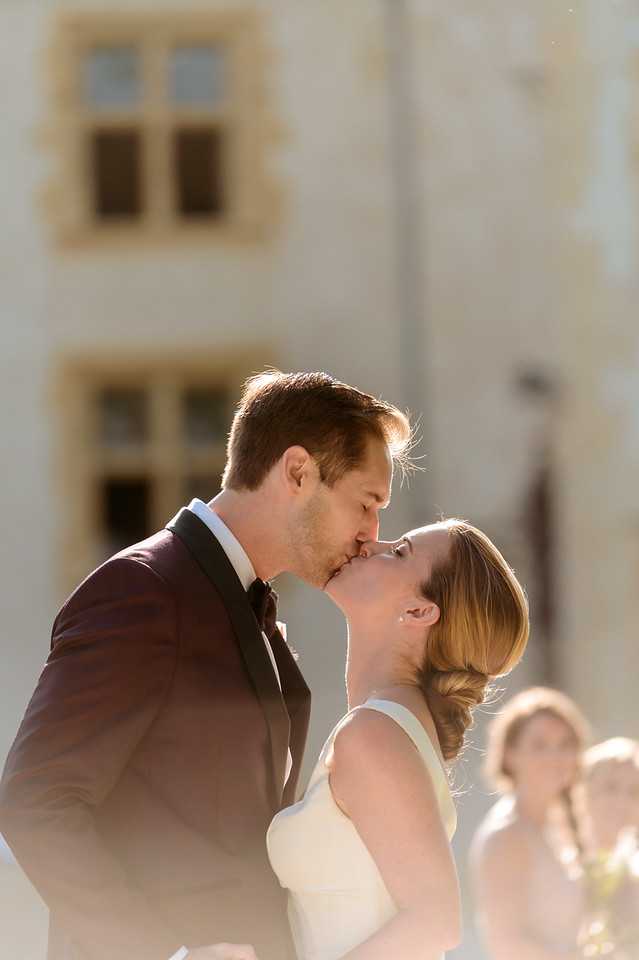 A couple shares their first kiss during an outdoor wedding ceremony, photographed in a tight portrait composition with shallow depth of field. The groom wears a deep burgundy tuxedo jacket with black lapels and a black bow tie, while the bride wears a sleeveless ivory fitted dress with her hair styled in an updo. The background shows the pale stone facade of what appears to be a French chateau with tall mullioned windows, softly blurred. At least two guests are visible but out of focus in the background lower right. Warm backlighting creates a sun-flare effect around the couple's heads.