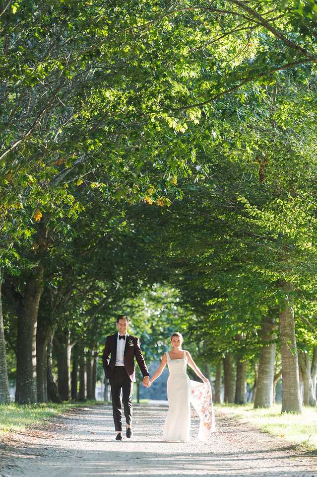 A couple walks hand-in-hand along a tree-lined gravel driveway in an outdoor portrait shot. The groom wears a dark burgundy tuxedo jacket with black trousers and a black bow tie, while the bride wears a fitted white sleeveless gown with a flowing train or overskirt featuring a floral print in pink and orange tones. The avenue of mature trees forms a natural canopy overhead, framing the couple along the full-length perspective of the path. The composition is a wide, centered portrait with the couple positioned in the middle distance, walking toward the camera.