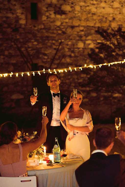 The couple is toasting with champagne flutes during an outdoor evening reception, standing at their sweetheart table surrounded by a small group of guests who raise their glasses in return. The setting is an outdoor courtyard or terrace against a rustic stone wall, with warm fairy lights strung overhead and candles on the table providing the primary lighting. The groom wears a dark tuxedo with a black bow tie, while the bride wears a strapless white dress with a white wrap or bolero jacket. The table displays a floral centerpiece with warm-toned blooms, candles, and a bottle of water, with a guest in a dusty pink dress visible in the foreground. The shot is a medium portrait taken at slight distance, capturing the warm, candlelit atmosphere of the nighttime reception.