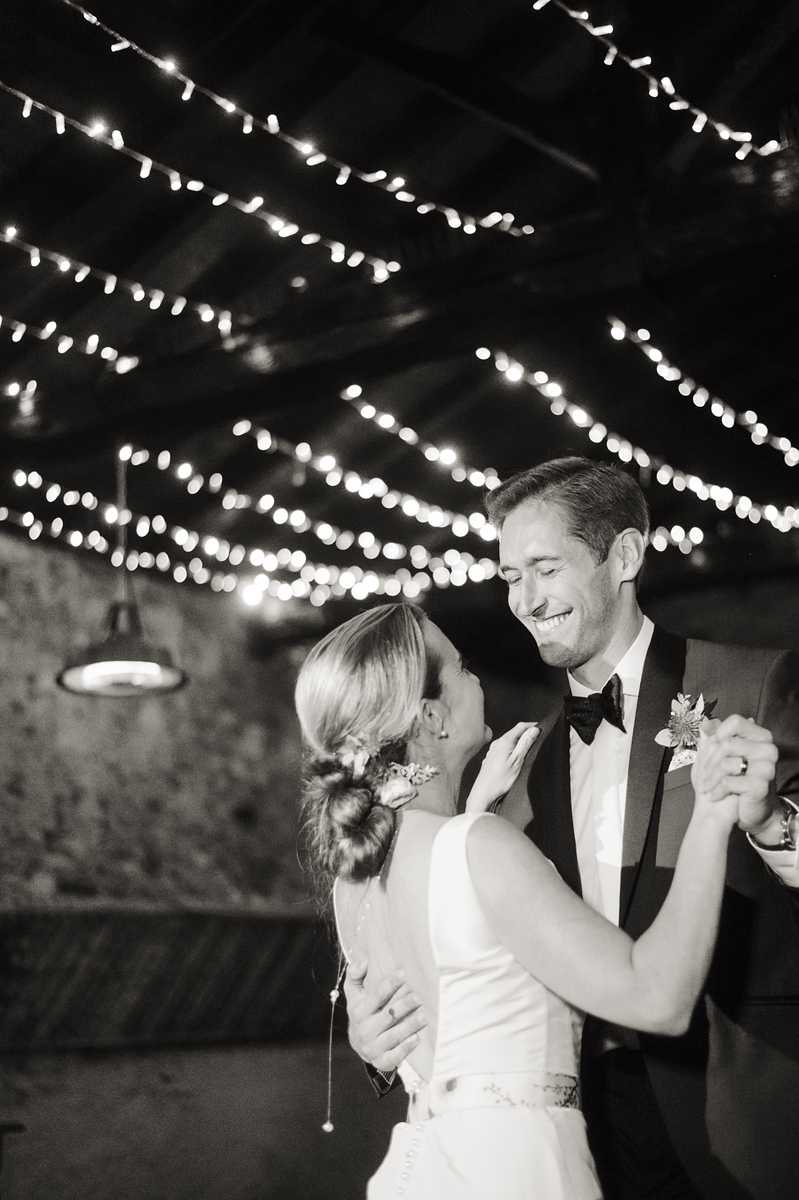 Black-and-white portrait-style shot of a couple sharing their first dance indoors at an evening reception. The bride wears a sleeveless fitted gown with a beaded belt detail and has her hair in a low twisted updo with small floral pins; the groom wears a dark tuxedo with a bow tie and a floral boutonniere on his lapel. Multiple strands of fairy lights are draped across a dark wood-beamed ceiling overhead, creating bright bokeh highlights against the dark background. The image has high contrast with deep shadows and bright highlights from the fairy lights, and a pendant lamp is visible on the left side of the frame.