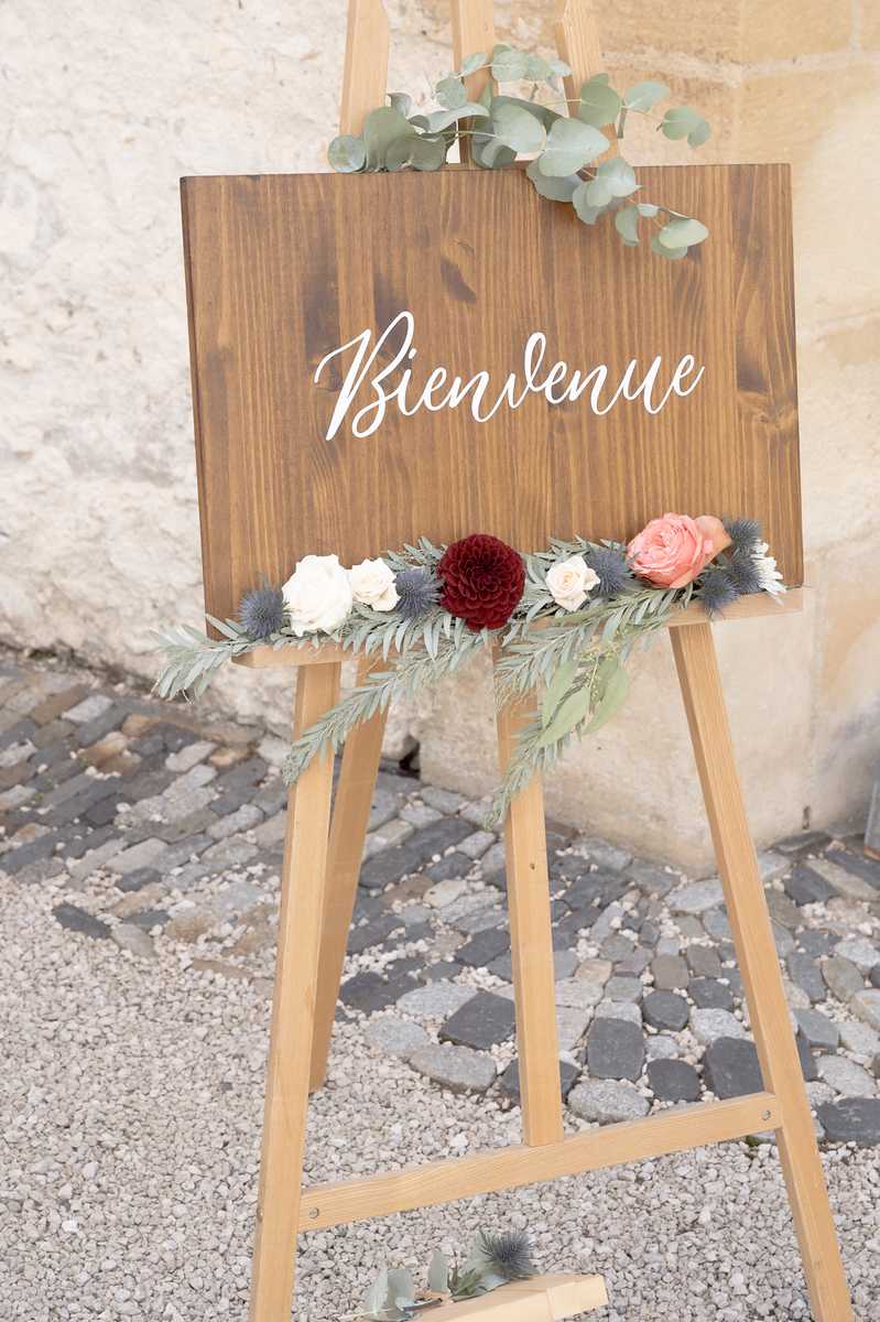 Close-up detail shot of a wedding welcome sign displayed on a natural wood easel, positioned outdoors on a cobblestone and gravel surface against a stone wall. The sign is a dark walnut-stained wooden board with 'Bienvenue' written in white script lettering. The bottom edge of the sign is decorated with a floral arrangement featuring a deep burgundy dahlia, cream roses, a coral garden rose, blue eryngium thistles, and eucalyptus and olive-leaf greenery, with additional eucalyptus sprigs placed at the top of the sign. The overall styling is rustic-classic with a warm, earthy color palette of burgundy, coral, cream, and dusty blue.