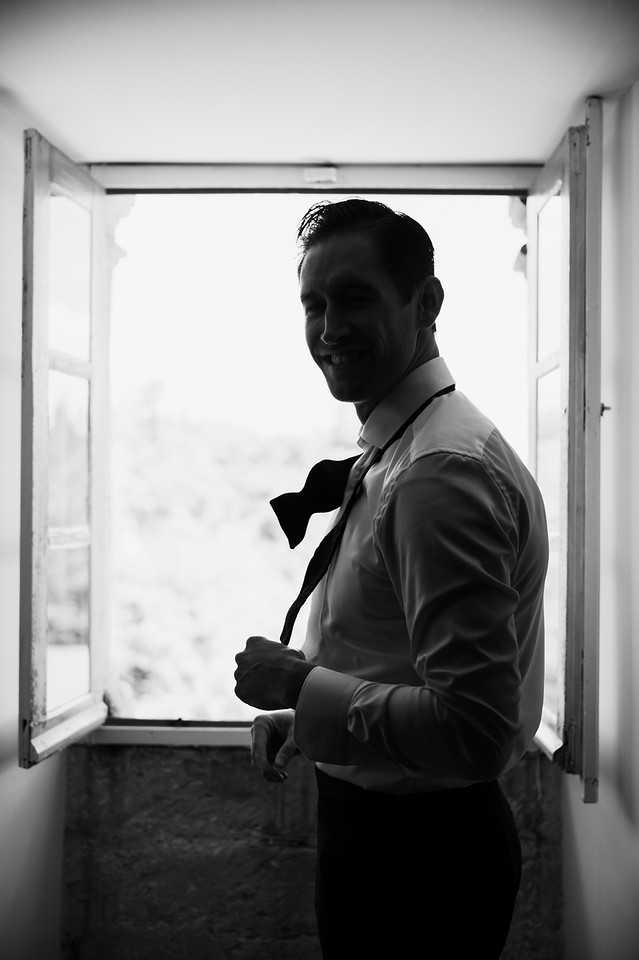 Black-and-white portrait of a groom getting ready indoors, standing in front of an open casement window that backlit his figure with bright light. He is wearing a white dress shirt and adjusting a bow tie that hangs loosely around his collar, smiling slightly downward. The high contrast between the bright window and the darker interior creates a semi-silhouette effect, with his facial features and shirt details still visible. The composition is a medium portrait shot taken from a slight low angle, emphasizing the mood of a private pre-ceremony getting-ready moment.