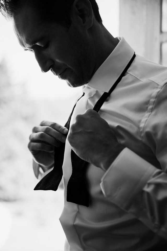 A black-and-white close-up portrait of a groom getting ready, captured as he adjusts a bow tie at his collar. He is wearing a white dress shirt with French cuffs and is looking downward with a slight smile. The image uses strong contrast with deep shadows on one side of his face and bright highlights from what appears to be a nearby window. The composition is a tight upper-body shot with a softly blurred background, emphasizing the getting-ready moment.