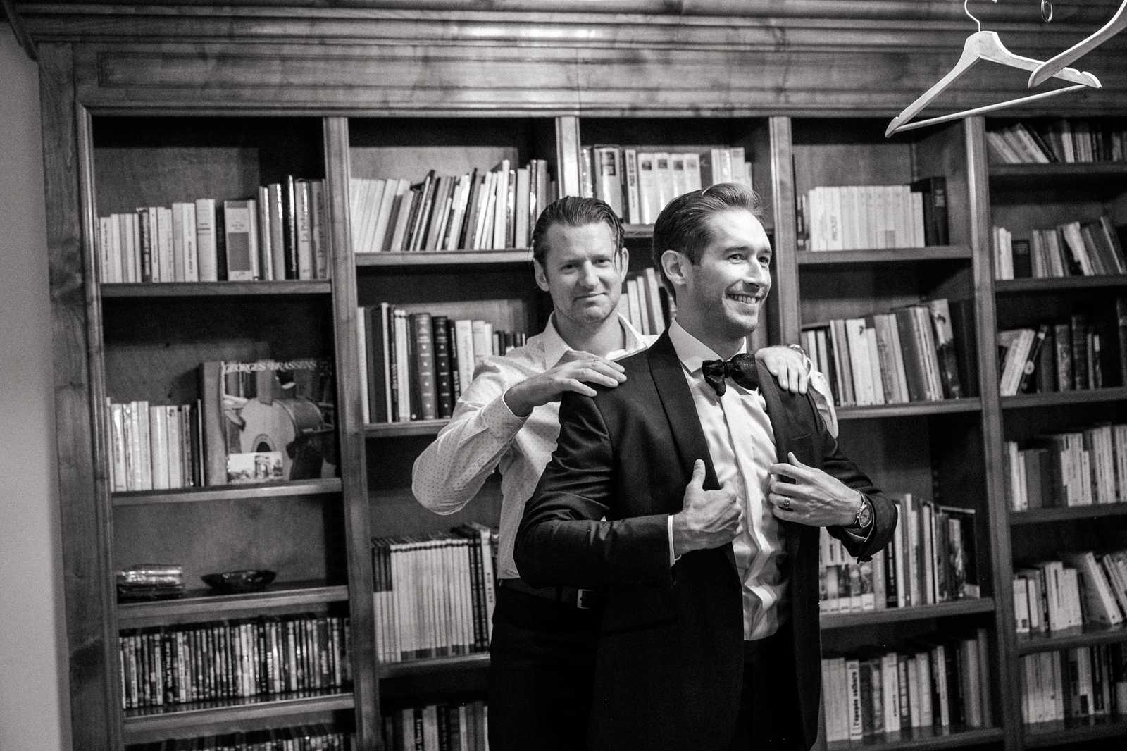 A black-and-white getting-ready shot of two men in a wood-panelled library room filled with books on floor-to-ceiling shelves. The groom, wearing a dark tuxedo jacket with a bow tie and white dress shirt, is smiling as a companion in a casual dress shirt helps adjust his jacket lapels from behind, with hands placed on his shoulders and chest. A white clothing hanger is visible in the upper right corner of the frame, and a Georges Brassens record or book cover is visible on one of the shelves. The image has strong contrast with deep shadows and bright highlights, shot at mid-range as a two-person portrait.