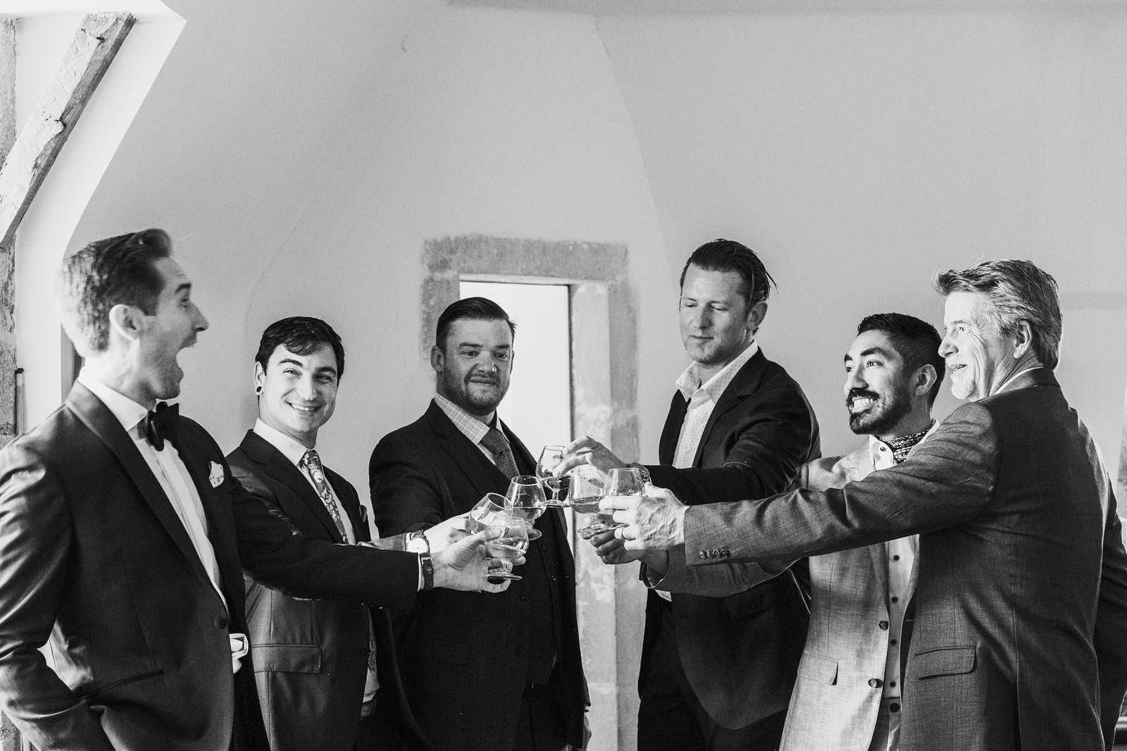 This black-and-white image shows six men raising glasses in a toast indoors, likely during a pre-ceremony getting-ready moment. The groom stands at the far left wearing a tuxedo with a black bow tie and pocket square, while the five other men — presumably groomsmen and family members — are dressed in a mix of dark suits with ties and sport jackets with open-collar shirts. The setting appears to be inside a rustic venue with arched ceilings, exposed timber beams, and whitewashed walls visible in the background. The image is a candid mid-shot with high contrast black-and-white tones, capturing the group laughing and smiling as they clink wine glasses together.