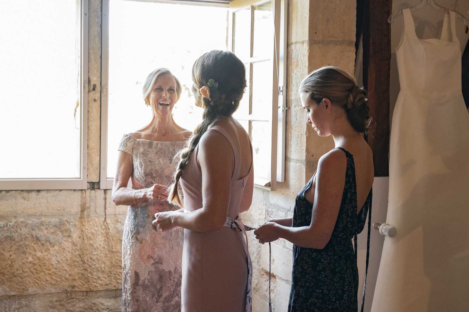 A getting-ready scene taking place indoors in a room with exposed stone walls, likely within a French chateau or historic venue. Three women are gathered near an open white-shuttered window: a woman in a floral-lace off-the-shoulder light pink-grey dress is smiling broadly, while a second woman with a braided updo adorned with a peach flower hair accessory and wearing a dusty mauve sleeveless dress stands with her back to the camera, and a third woman in a dark navy floral slip dress appears to be tying or fastening the back of the center woman's dress. A white wedding gown hangs on a hanger against the stone wall in the upper right corner of the frame. The shot is a medium-wide portrait taken from behind the central figure, with natural backlight streaming through the open window.