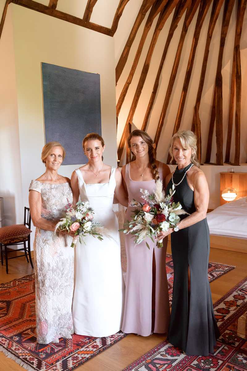 A getting-ready or pre-ceremony portrait of four women standing together indoors, including the bride and three female guests or family members. The bride wears a white square-neck satin gown, while the other women wear a silver-grey lace floor-length dress, a blush pink satin slip dress, and a dark charcoal halter-neck gown respectively. Three of the four women hold bouquets featuring blush garden roses, burgundy dahlias, white blooms, pampas grass, thistles, and mixed greenery in an organic, loosely arranged style. The setting is a rustic interior room with exposed dark timber beams radiating from the ceiling, whitewashed walls, a large abstract blue-grey canvas painting, a patterned red and navy kilim rug on the hardwood floor, and a warmly lit wooden bedside lamp visible in the background. The overall styling aesthetic mixes rustic architectural features with a modern, minimalist dress palette. Full-length group portrait shot.
