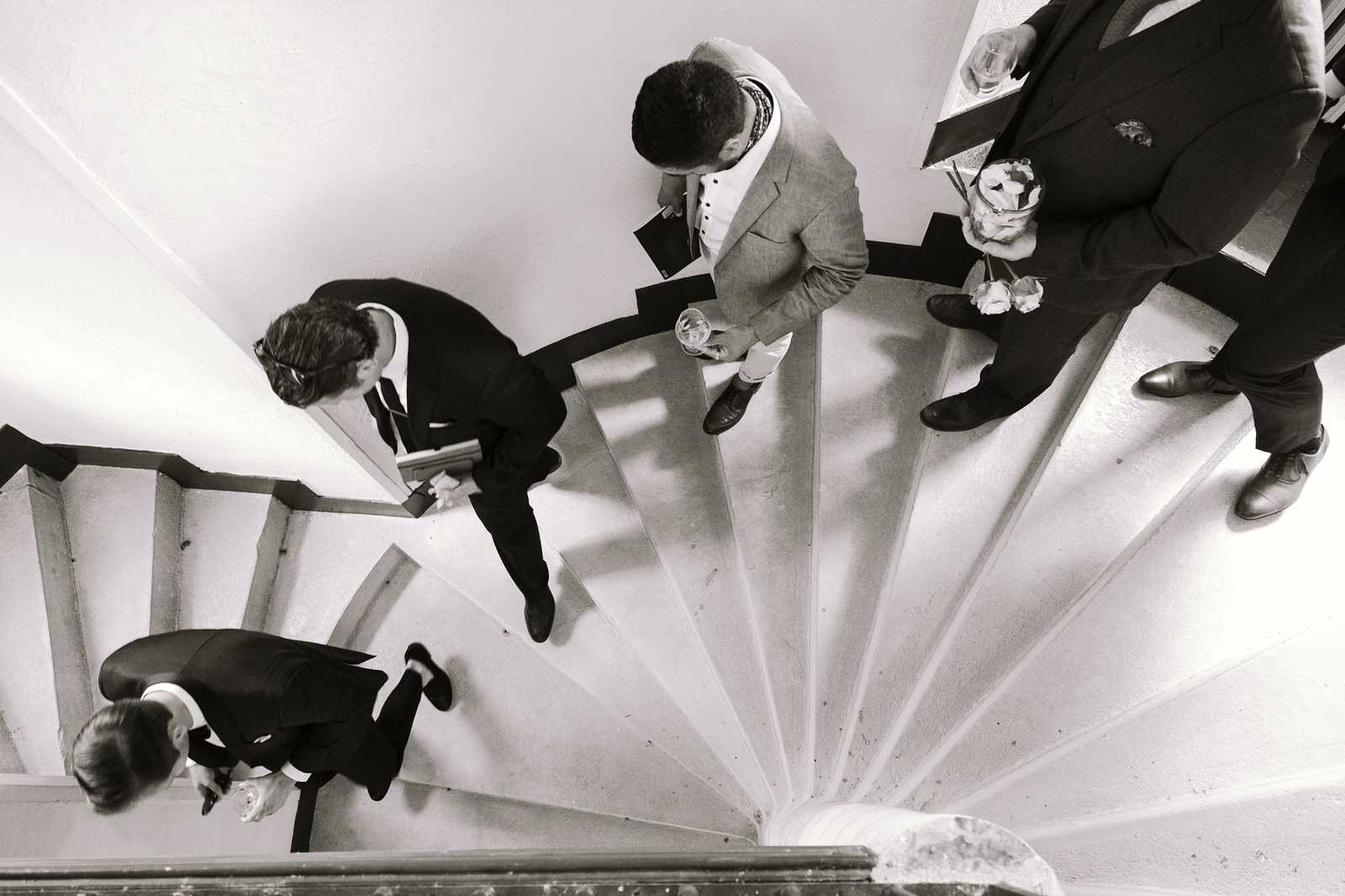 This black-and-white image is shot from directly overhead, looking down onto a stone spiral staircase where four formally dressed wedding guests are gathered. The composition emphasizes the geometric spiral pattern of the staircase steps, with strong contrast between the bright white treads and dark shadows. Three of the guests appear to be in dark suits, while one wears a light-colored (likely beige or grey) suit; at least one person is holding a drink and another appears to be holding white flowers, likely boutonnieres or a small bouquet. The overhead aerial angle creates a graphic, documentary-style shot typical of candid wedding reportage photography.