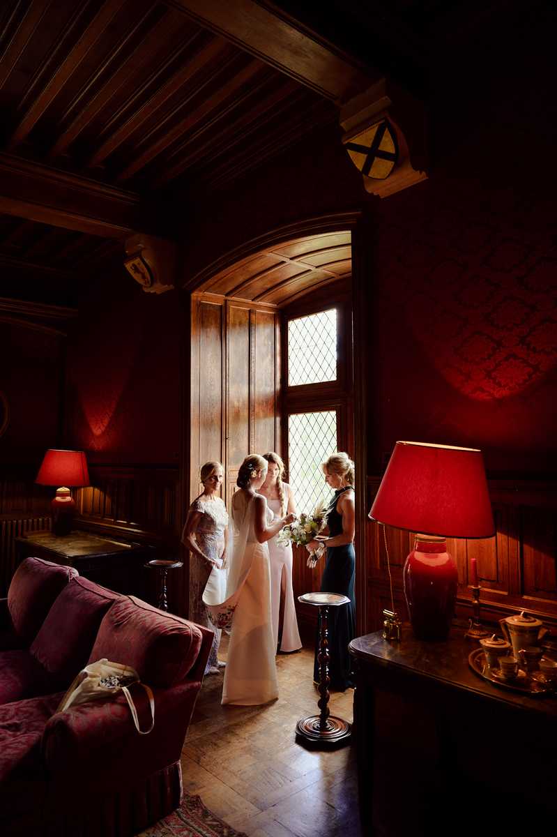 A getting-ready or pre-ceremony moment inside a richly decorated chateau room, where a bride in a sleek ivory satin gown with a veil holds a white and green bouquet while interacting with two women — one in a floral lace dress and one in a dark navy fitted dress. The room features dark wood paneling, a coffered wooden ceiling, leaded diamond-pane arched windows providing backlighting, deep red damask wallpaper, two red-shaded table lamps casting warm ambient light, a burgundy velvet sofa, parquet flooring, and antique furnishings including a sideboard with a tea service. The overall color palette is deep crimson, mahogany, and gold with the bride's white gown as the focal point against the window light. Wide portrait shot taken from a low-to-mid angle, emphasizing the dramatic dark interior contrasted with the natural window light. Potential venue feature image.