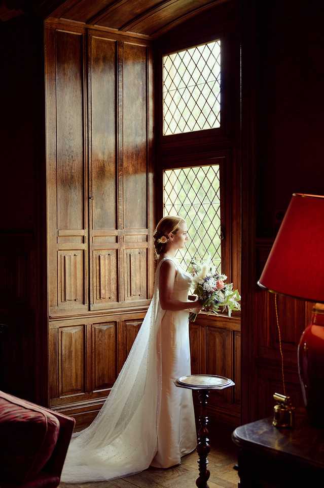A bride stands alone in a richly wood-paneled interior room, looking out through a tall leaded diamond-pane window. She wears a sleek ivory gown with a long train and has her hair pinned up with a small floral hair accessory. She holds a loose, textured bouquet featuring coral, dusty blue, and white blooms with greenery. The room is furnished with a dark wood pedestal side table, a deep red lampshade with a gold base, and what appears to be a burgundy upholstered chair, giving the space a classic, manor-house feel. The image is a full-length portrait shot, lit primarily by natural light from the window, with warm, moody shadows filling the surrounding interior.